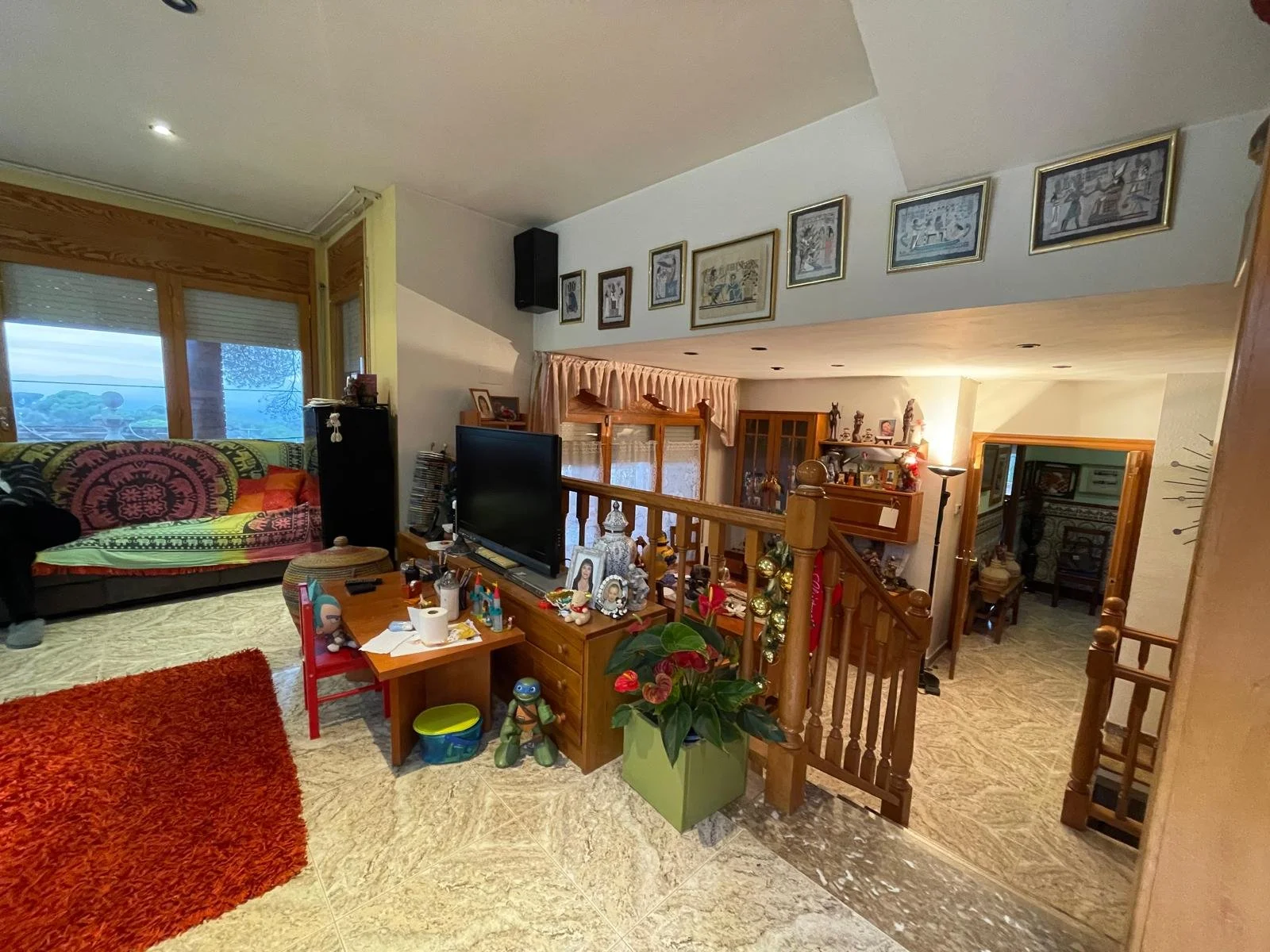 Sala de estar con sofá cubierto con manta colorida, televisión, mesa con juguetes y objetos decorativos, plantas, escaleras de madera, y varios cuadros en la pared.