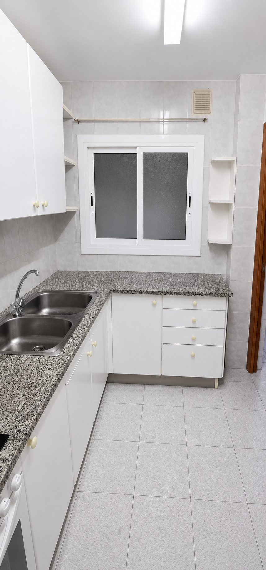 Cocina pequeña con azulejos blancos, encimera de granito, fregadero de doble cubeta, armarios blancos con pomo amarillo, ventana con vidrio esmerilado y estantes en la pared.