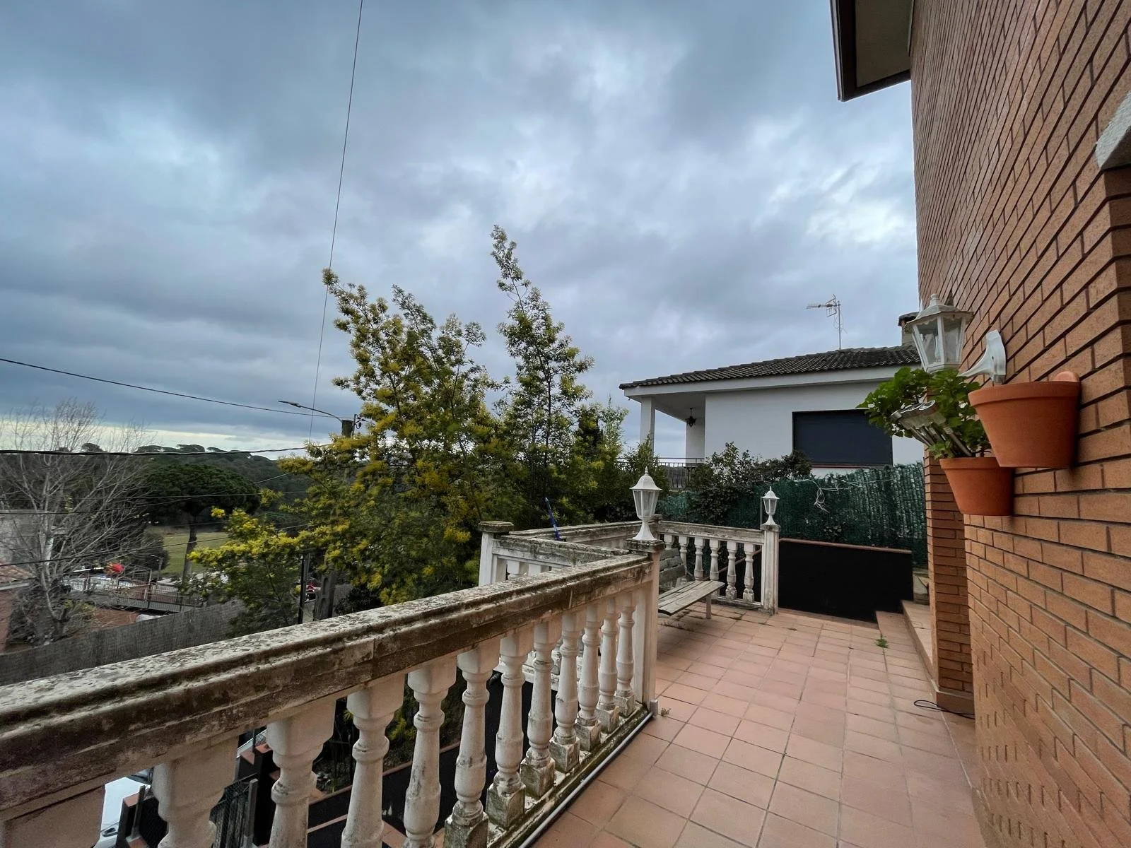 Vista de una terraza con barandal de cemento, varias plantas en macetas en la pared de ladrillos, un banco de madera, y un cielo nublado con árboles y casas al fondo.