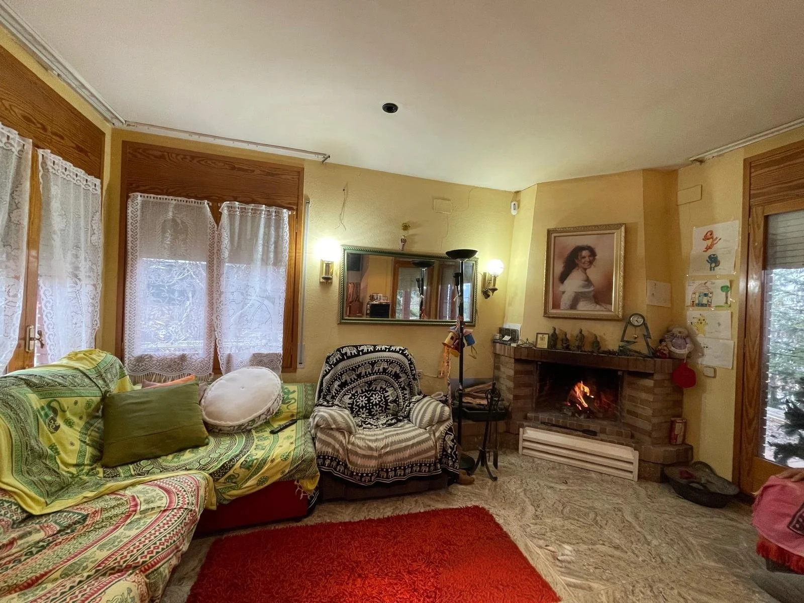 Sala de estar con sillón y sofá cubierto con mantas de colores, una chimenea encendida, un espejo grande en la pared, cuadros infantiles en la pared, cortinas de encaje, muchas decoraciones y una alfombra roja.