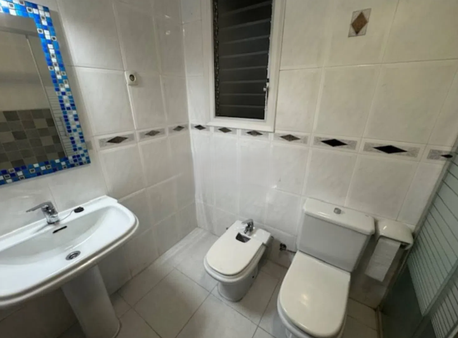Baño con lavabo, inodoro y bidé, paredes con azulejos blancos y techo con ventana con cortina de persiana, espejo y radiador.