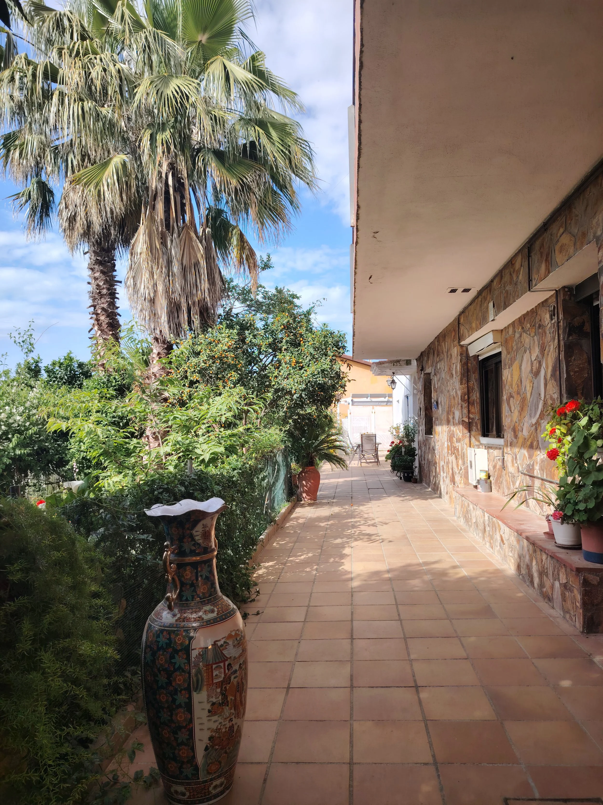 Un patio con plantas verdes y flores, una mesa y sillas en el fondo, paredes de piedra y un techo blanco
