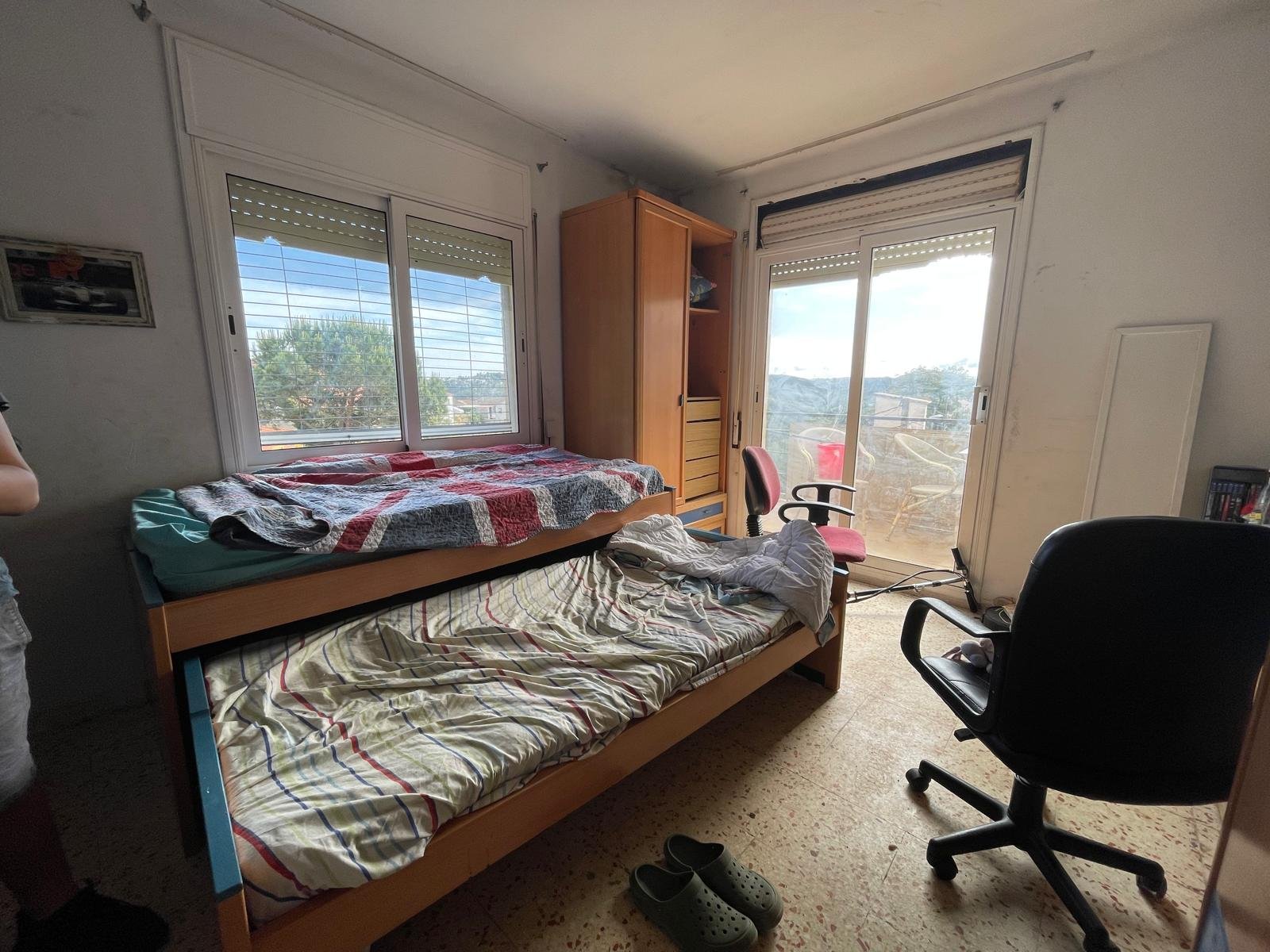 Una habitación desordenada con una cama sencilla cubierto con ropa y mantas, hay otra cama encima de la primera con una manta de cuadros, dos sillas, una mesa de madera, una ventana grande, una puerta corrediza de balcón, y un armario de madera.