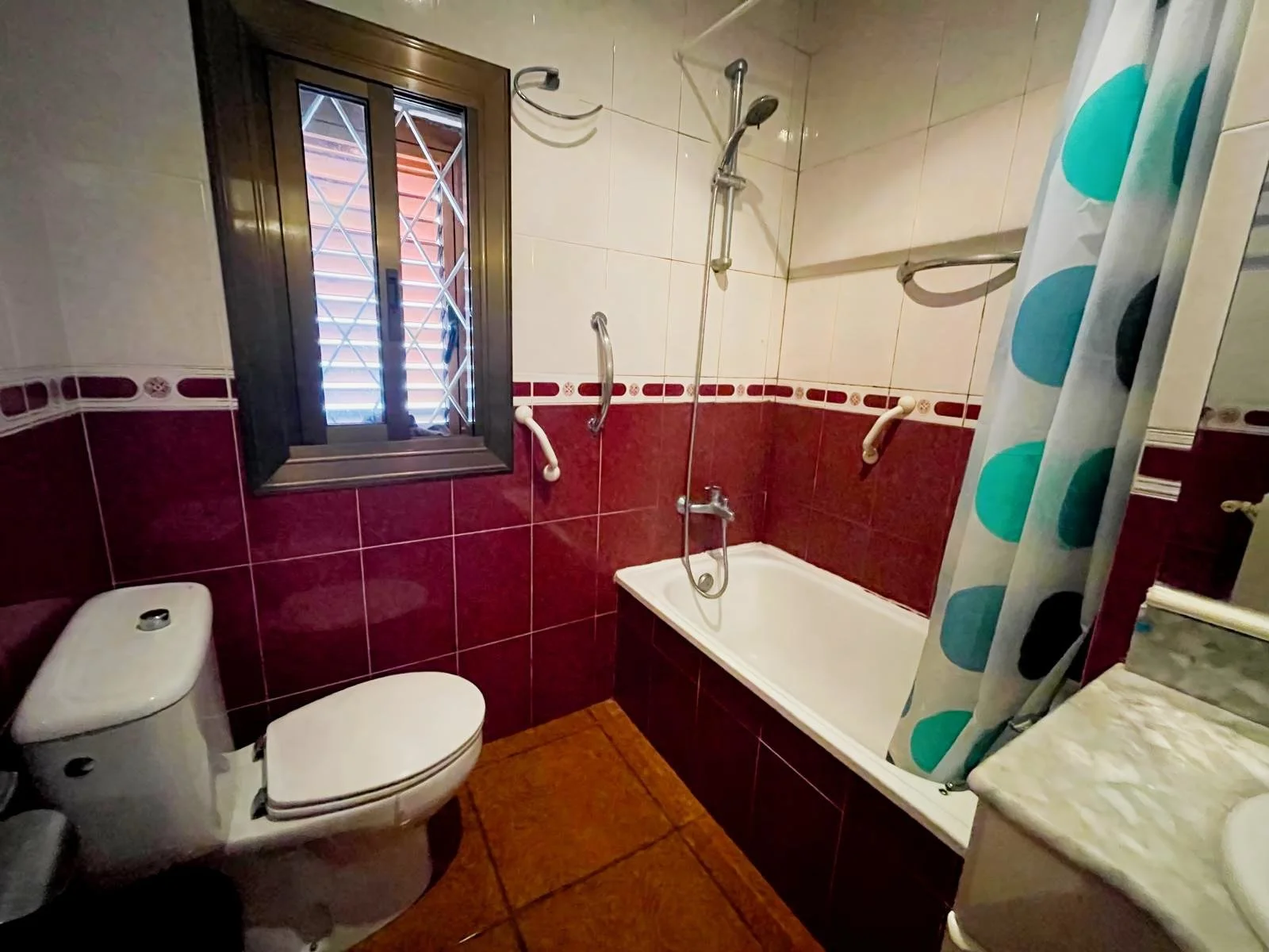 Baño con taza, ducha, bañera, y ventana con reja.