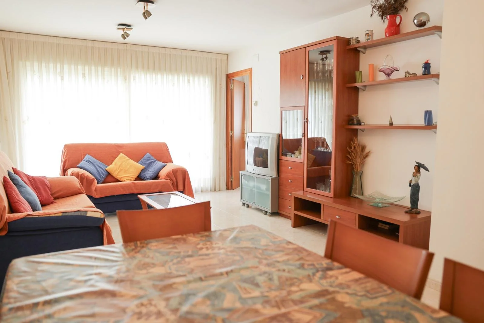 Sala de estar con sofás naranjas y cojines de diferentes colores, mesa de comedor con mantel, televisión antigua en carrito, armario de madera con espejo, estanterías con decoraciones, cortinas blancas y luz natural.