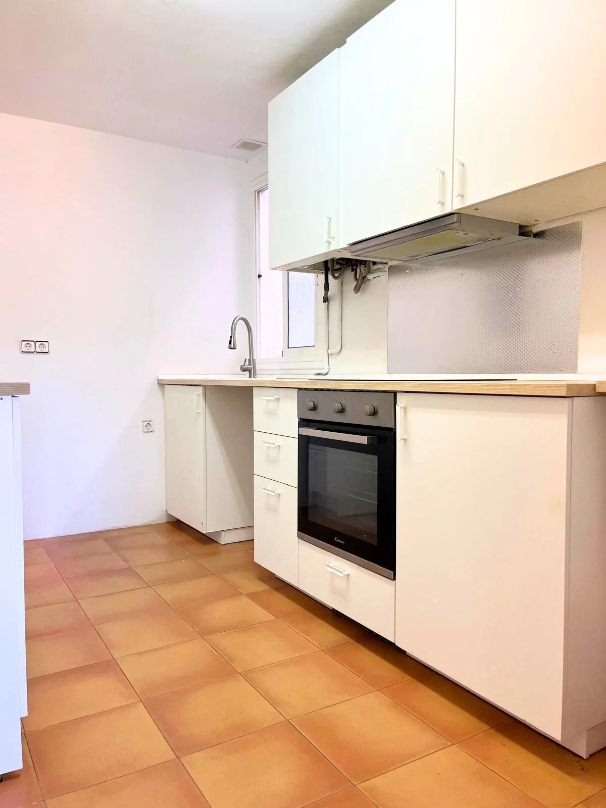Cocina moderna con gabinetes blancos, horno de wall empotrado, fregadero con grifo, ventana pequeña, suelo de baldosas terracota.
