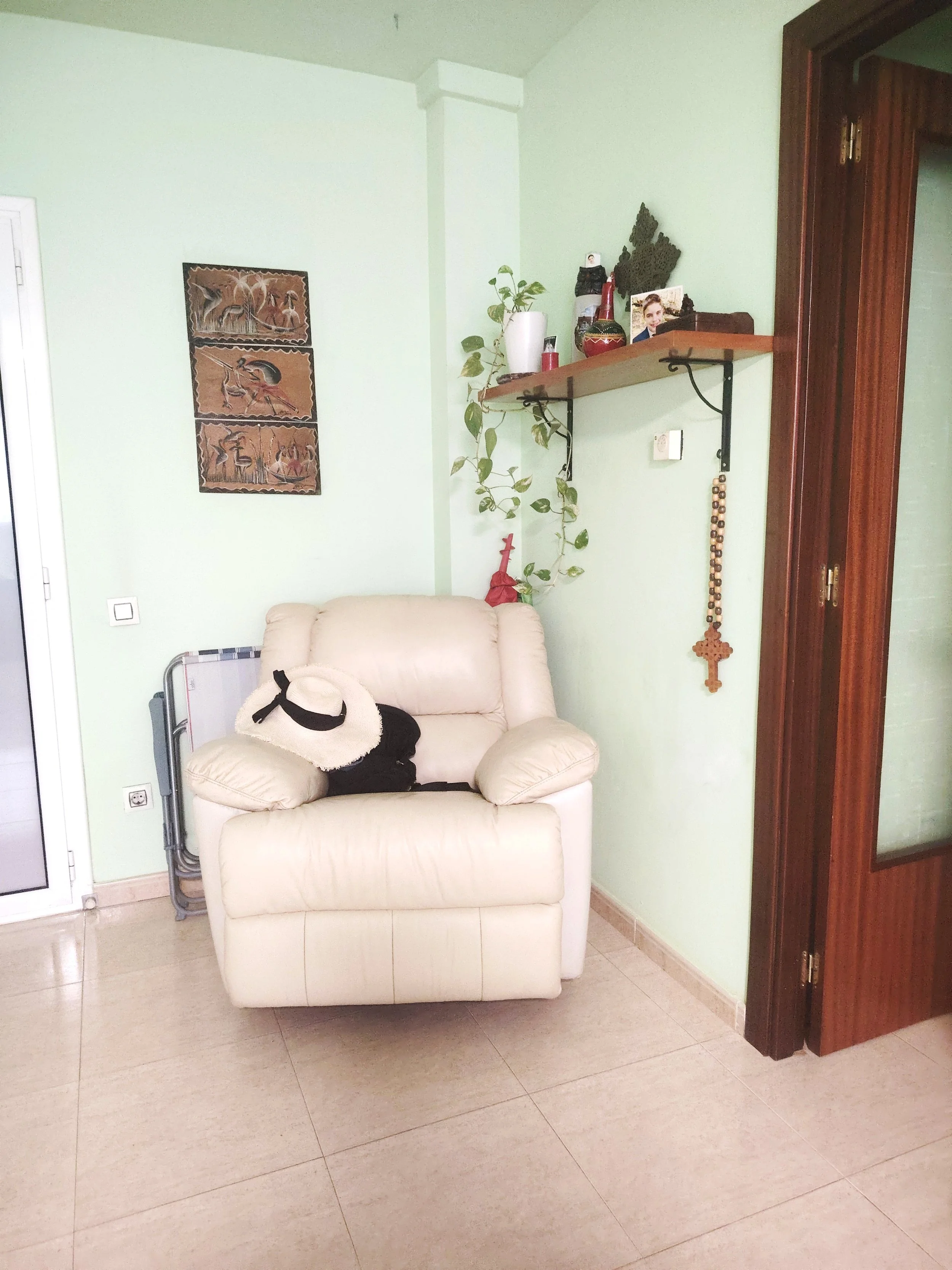 Sala con sillón beige, sombrero, estantería con plantas y objetos decorativos, cruz de rosario, cuadro con aves, pared de color verde claro y puerta de madera