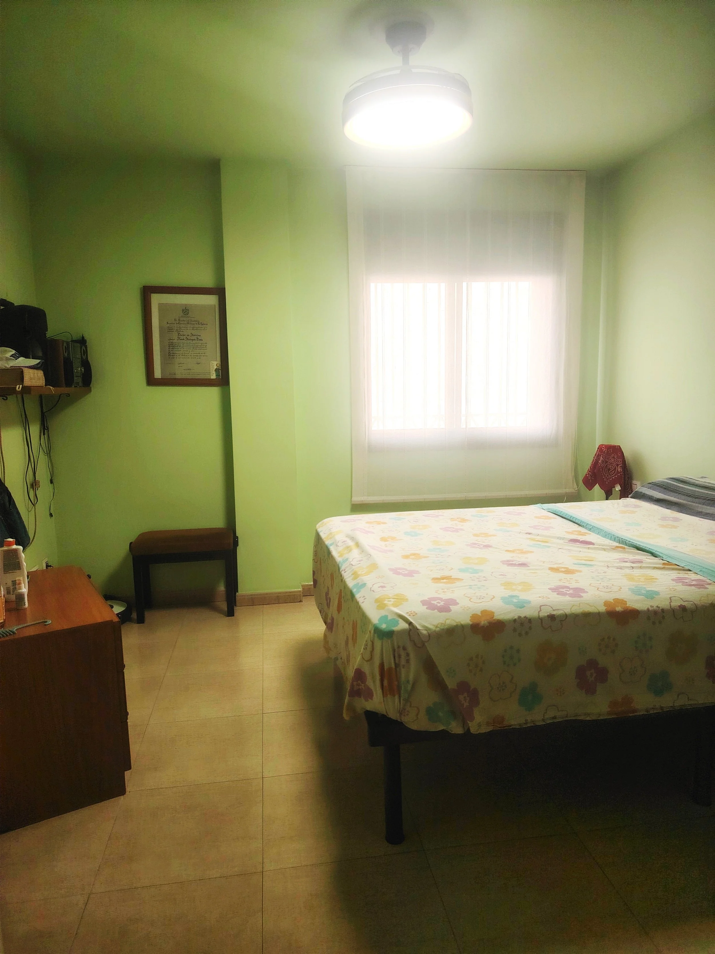 Habitación con paredes verdes, una cama con sábanas de colores con flores, una ventana con cortinas blancas, una lámpara en el techo y algunos muebles