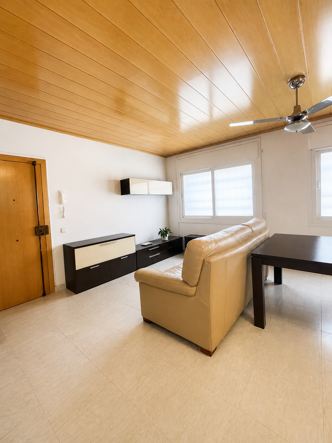 Sala de estar con techo de madera, sillón beige, mesa negra, ventanas con persianas, y un mueble de pared con plantas.