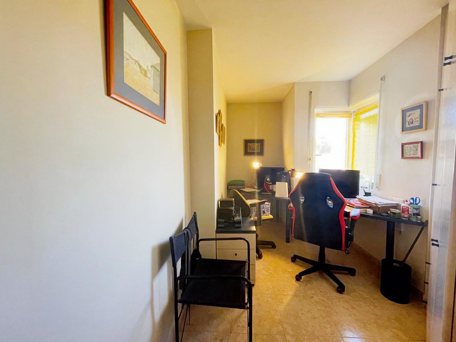 Oficina con dos escritorios, varias sillas, una ventana grande con cortinas y varios cuadros en las paredes.