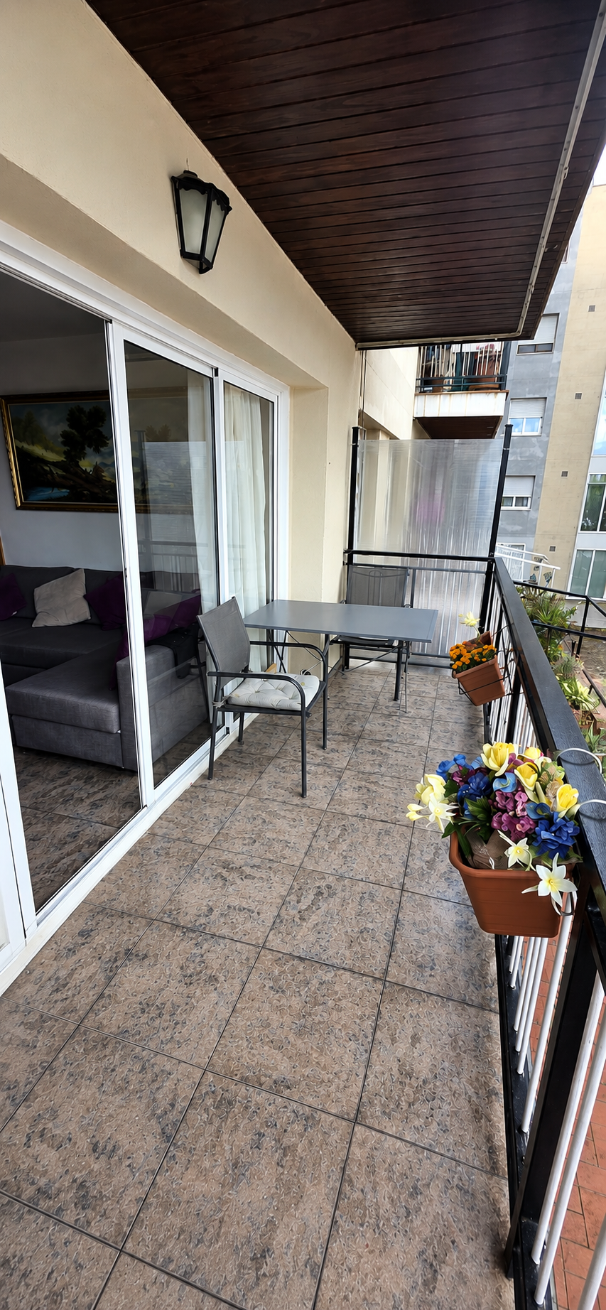 Balcón de apartamento con mesa y sillas, plantas con flores en macetas y vista a edificios vecinos.