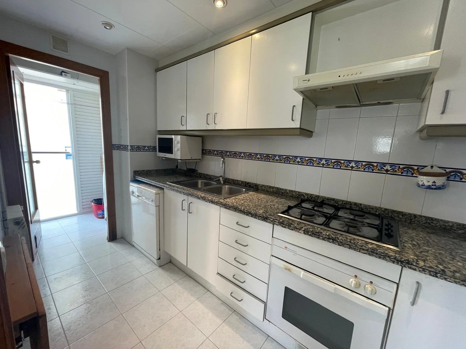 Cocina con gabinetes blancos, encimera de granito, estufa de gas integrada, horno, campana extractora, fregadero doble y microondas, con puerta al balcón al fondo.