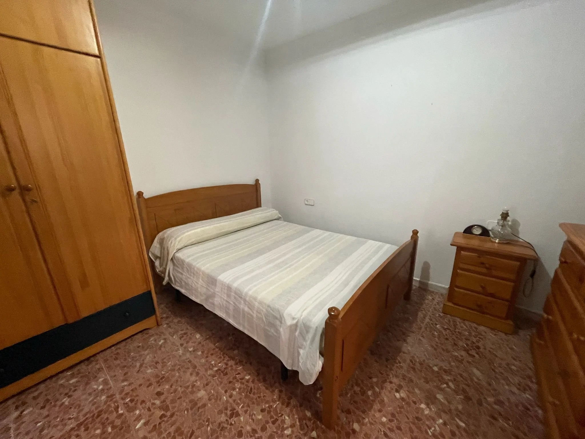 Habitación con cama doble, armario de madera, mesita de noche con reloj y botella, y suelo de azulejo