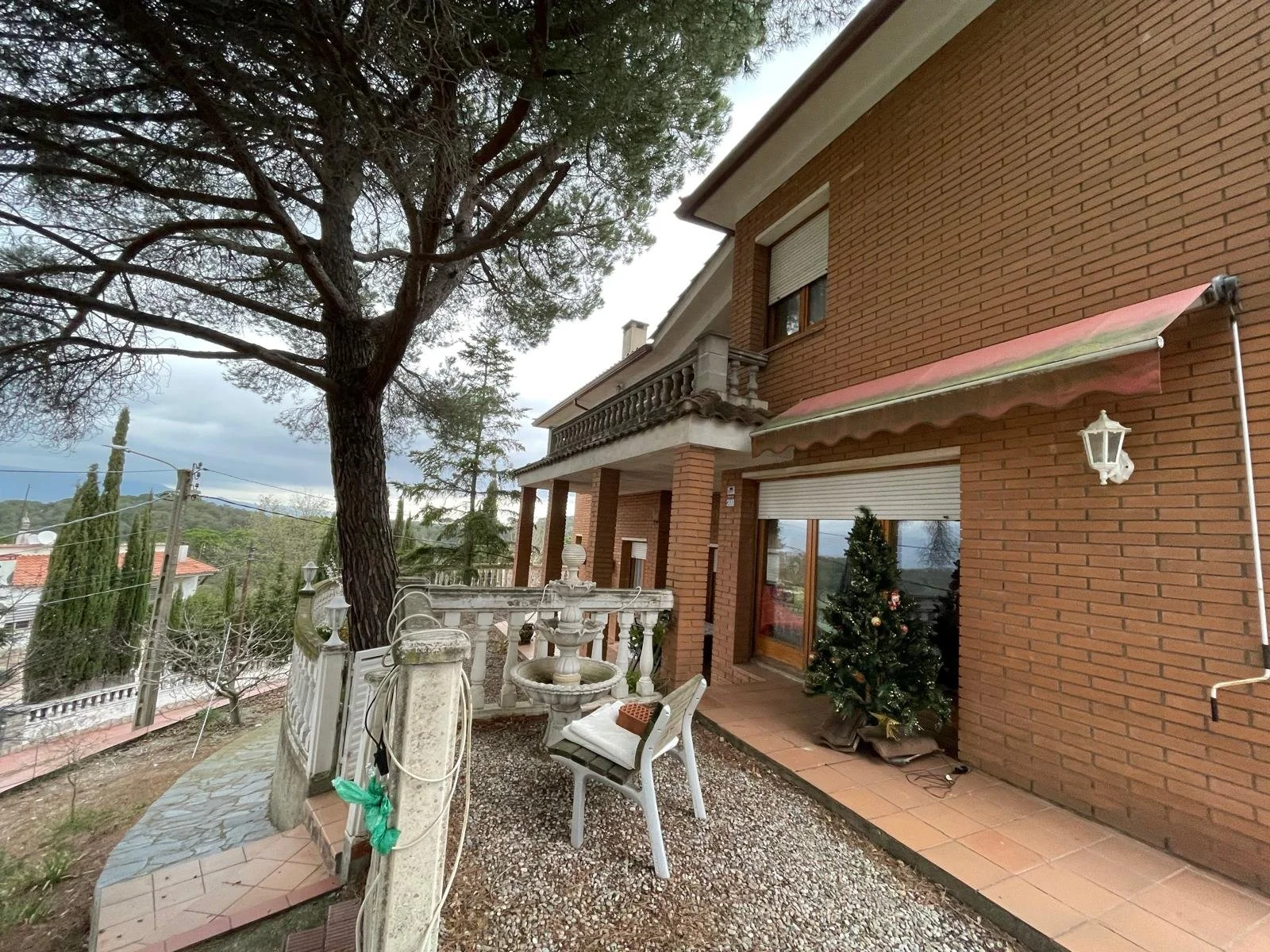 Fachada de una casa de ladrillo con un árbol grande y un balcón. En el patio hay una pequeña planta de Navidad decorada, una banca y una fuente. El cielo está nublado.