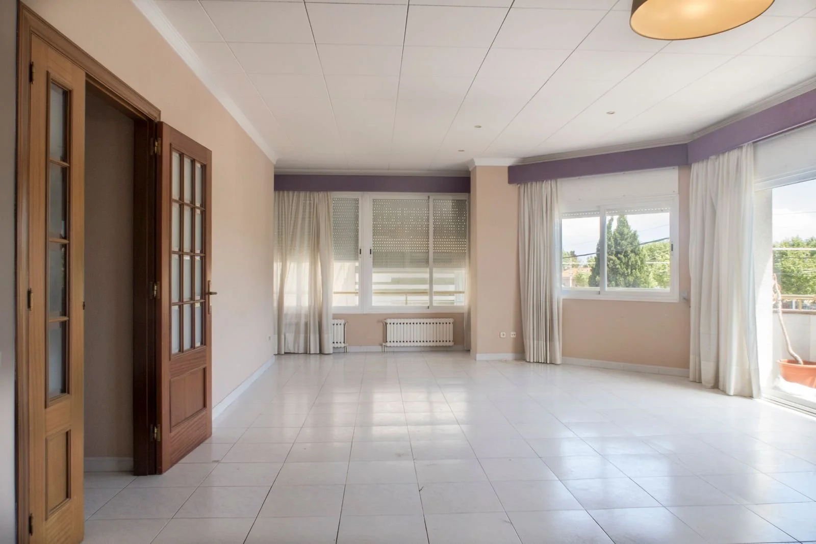Sala de estar con puertas de madera a la izquierda y grandes ventanas con cortinas blancas que dejan pasar luz natural.
