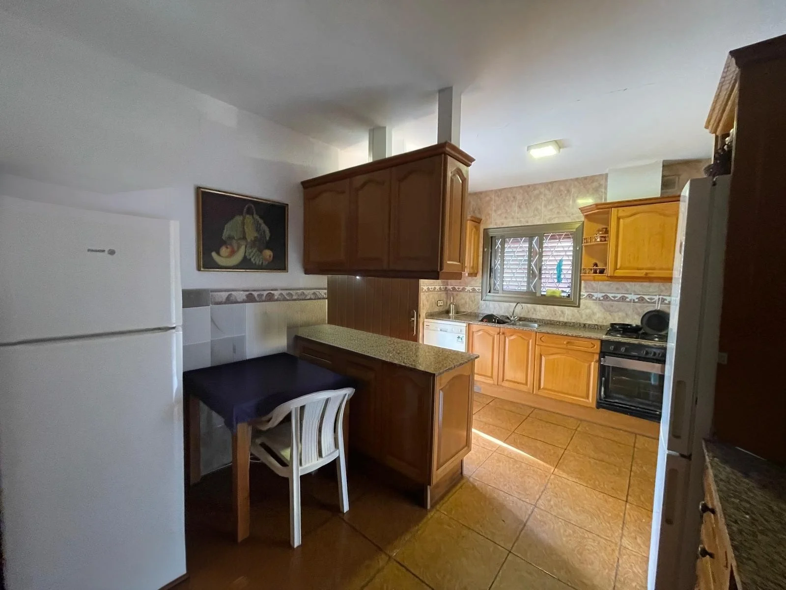 Cocina con electrodomésticos y muebles de madera, azulejos en paredes y piso, ventana con persianas, y mesa con sillas.