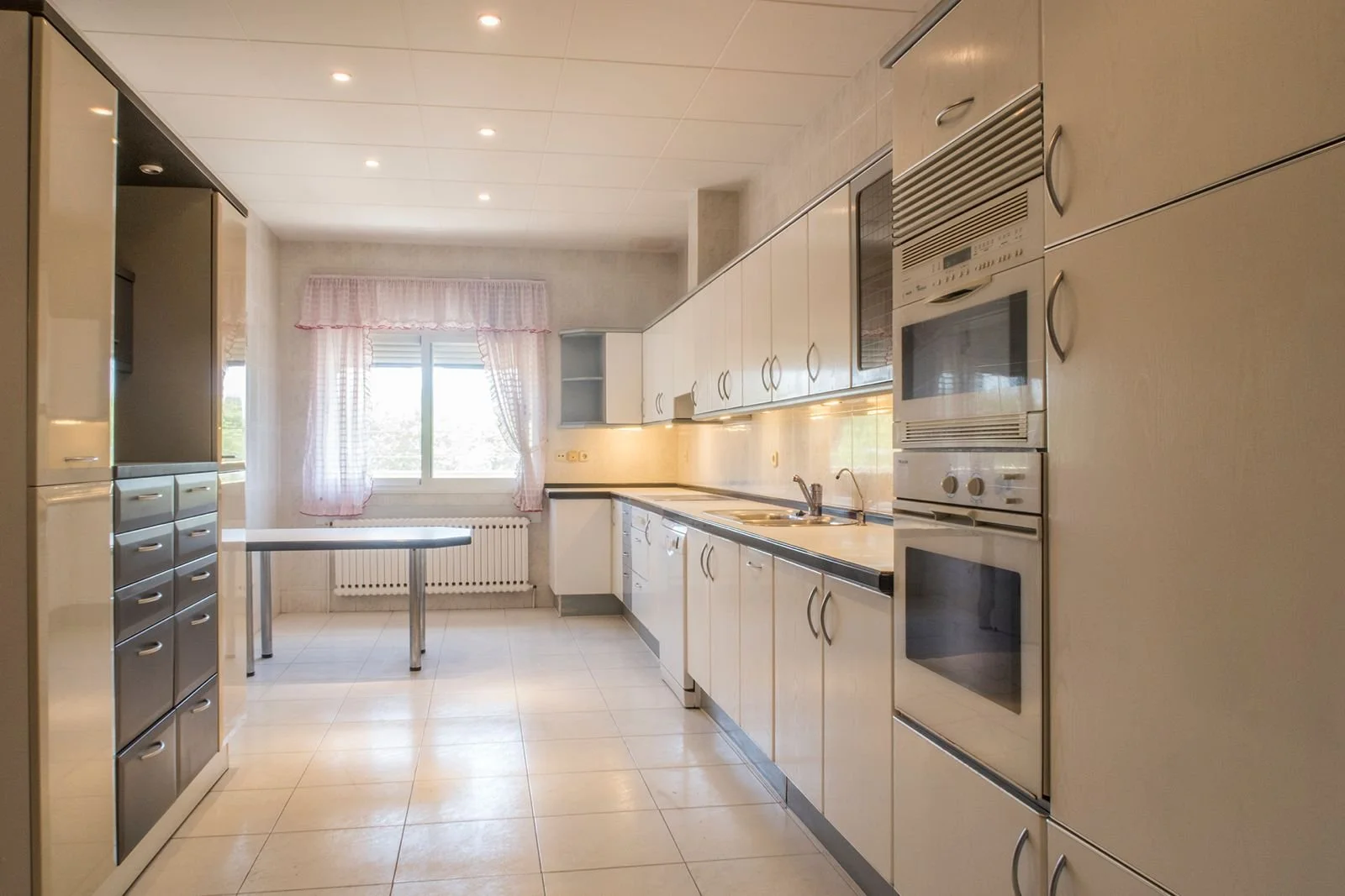 Cocina con gabinetes blancos, electrodomésticos empotrados, ventana con cortinas rosas, mesa pequeña y piso de baldosas beige.