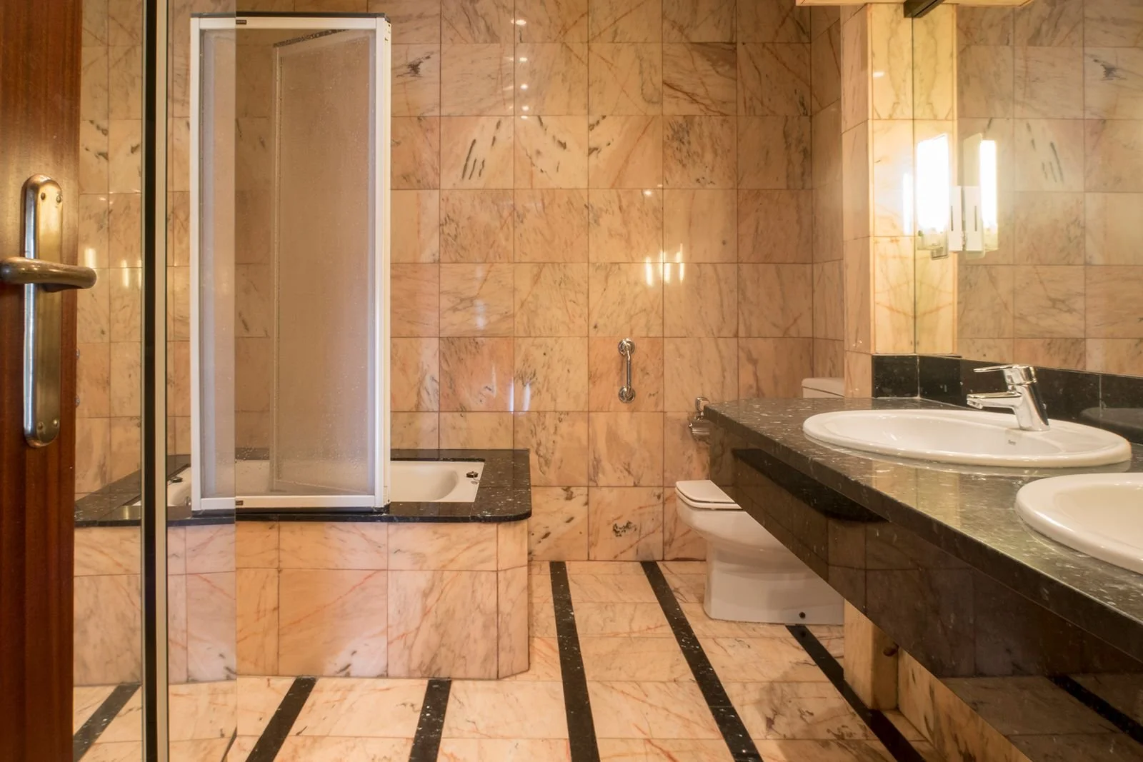 Baño con paredes y piso de mármol beige con vetas doradas y negras, ducha con puerta de acrílico, lavabo doble con encimera de granito negro, espejos con luces, inodoro y barras de apoyo.