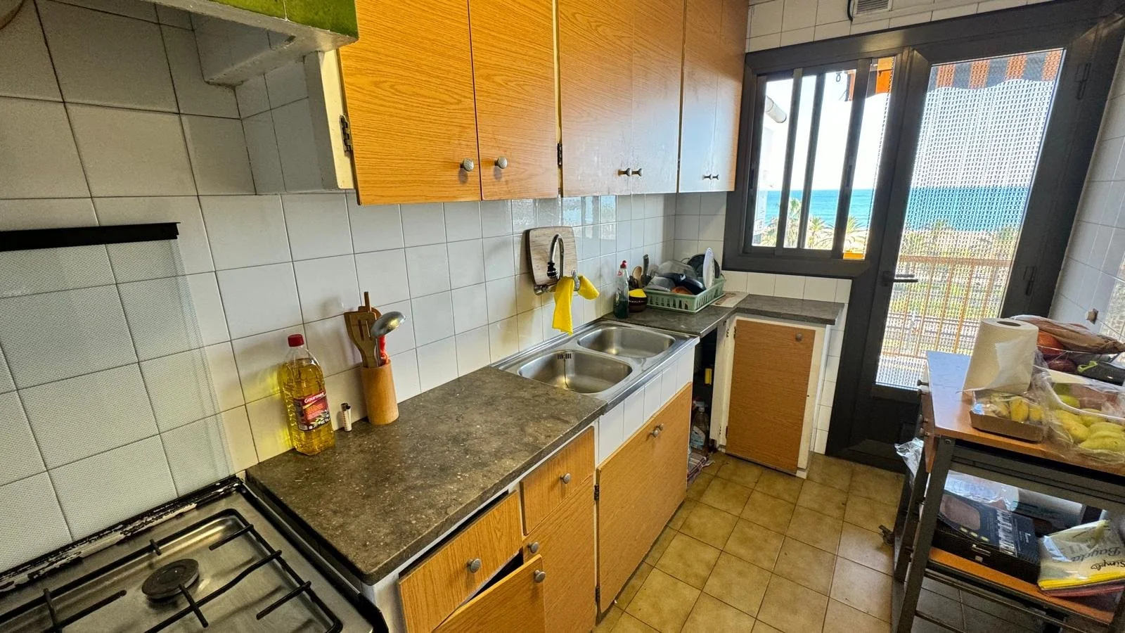 Cocina de apartamento con vista al mar, muebles de madera y encimeras de piedra, con utensilios y comida en preparación, ventana y puerta de balcón con vista al mar y palmeras.