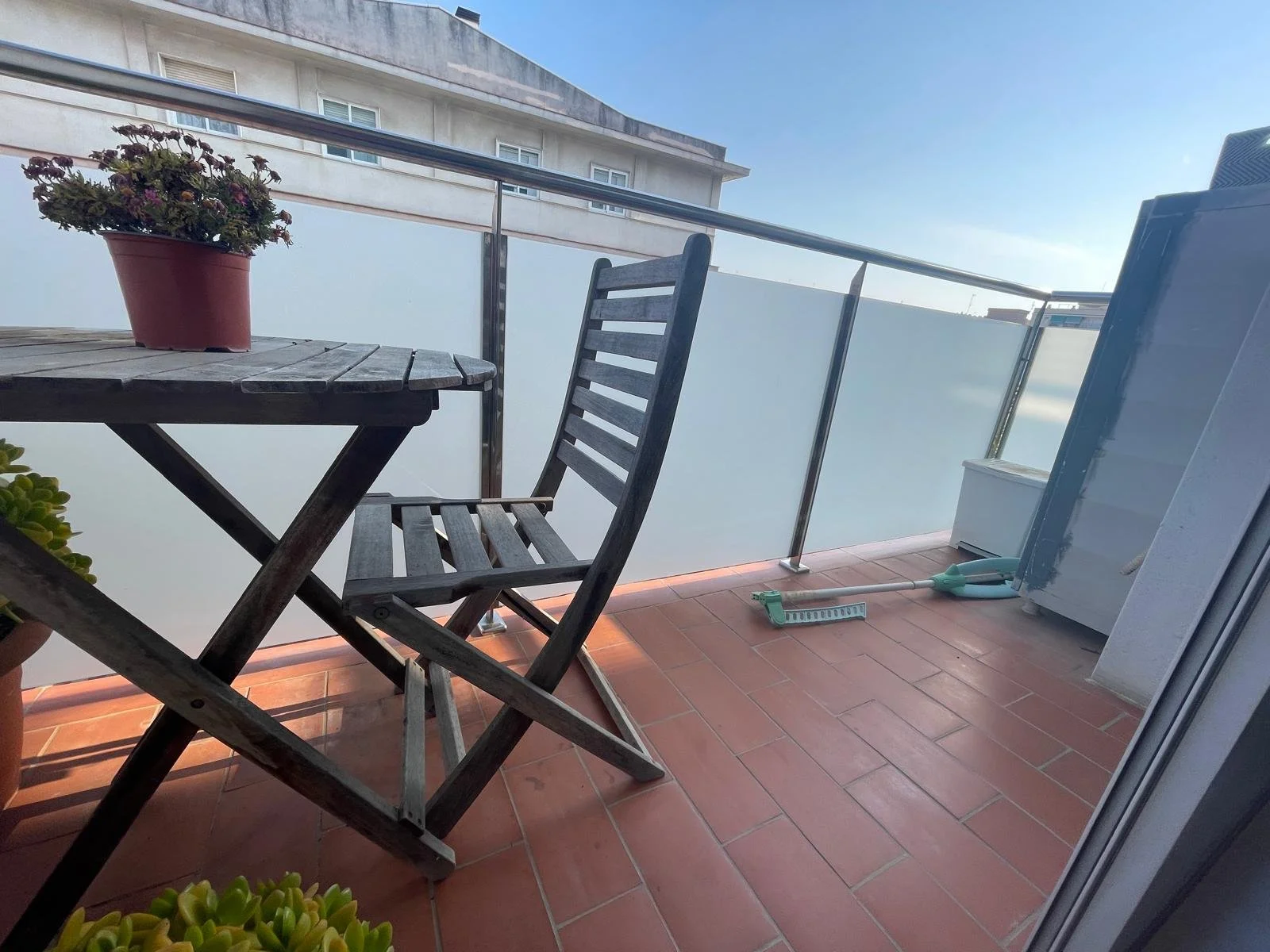 Balcony con mesa de madera y silla, plantas en las esquinas, trapeador y escoba en el piso, y vista de edificio y cielo azul.