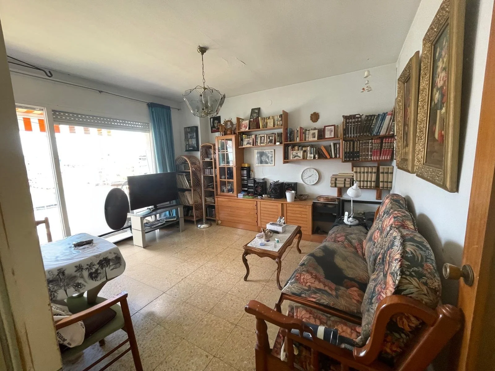 Sala de estar con sofá, mesa de café, estanterías con libros y objetos, televisor, silla, lámpara de escritorio, cuadros en las paredes y ventana con cortinas azules.