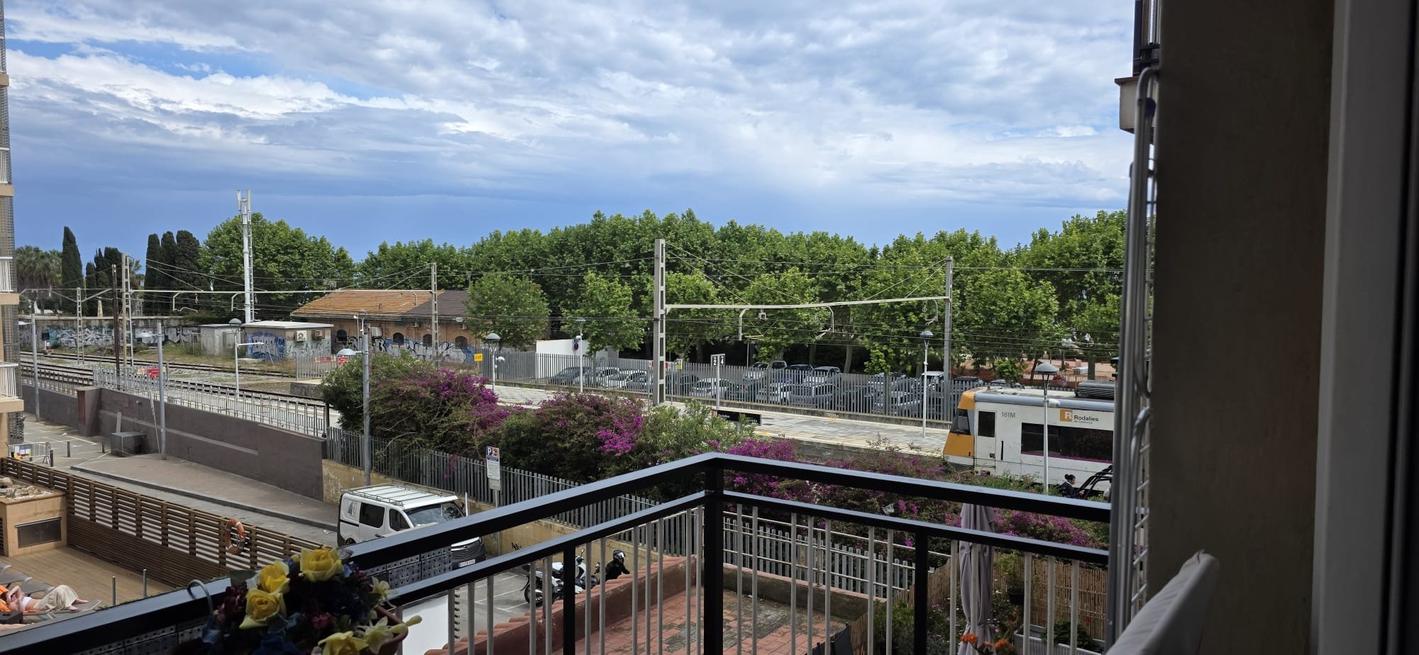 Vista de la calle desde un balcón, con árboles verdes y cielo nublado, cerca de un ferrocarril y transporte público, hay vehículos y personas en la calle.