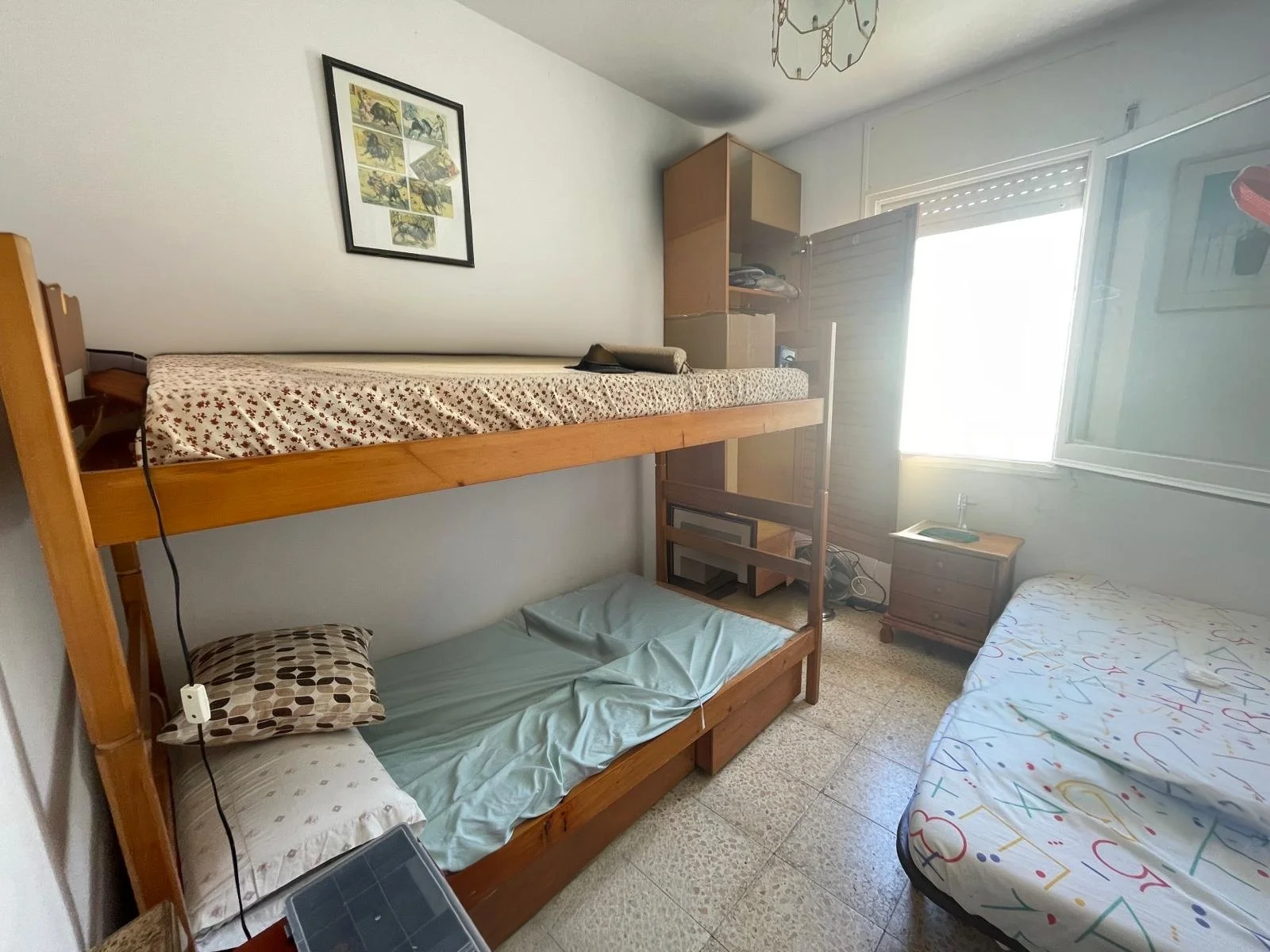 Dormitorio con literas de madera, una cama individual con sábanas blancas con dibujos de números, una pequeña mesa de noche, una ventana abierta y un armario junto a la ventana.