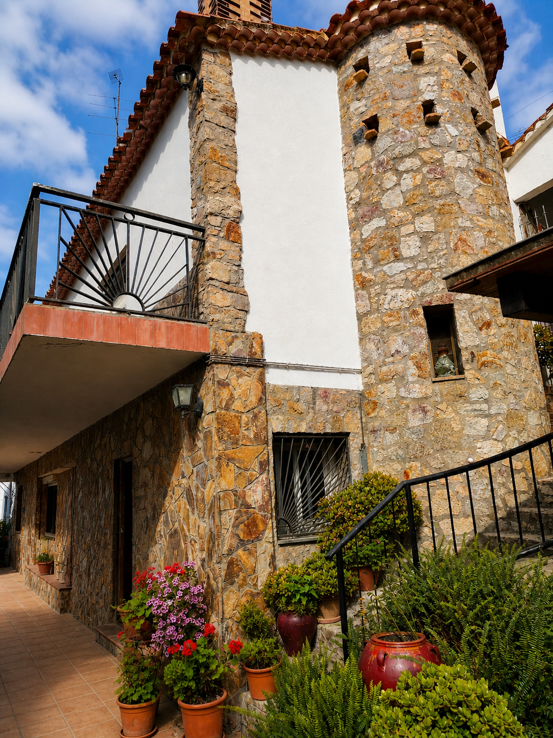 Casa con paredes de piedra y ventanas con rejas, barda de jardín con macetas de flores y plantas verdes, escalera con barandal, balcón con barandal de hierro.