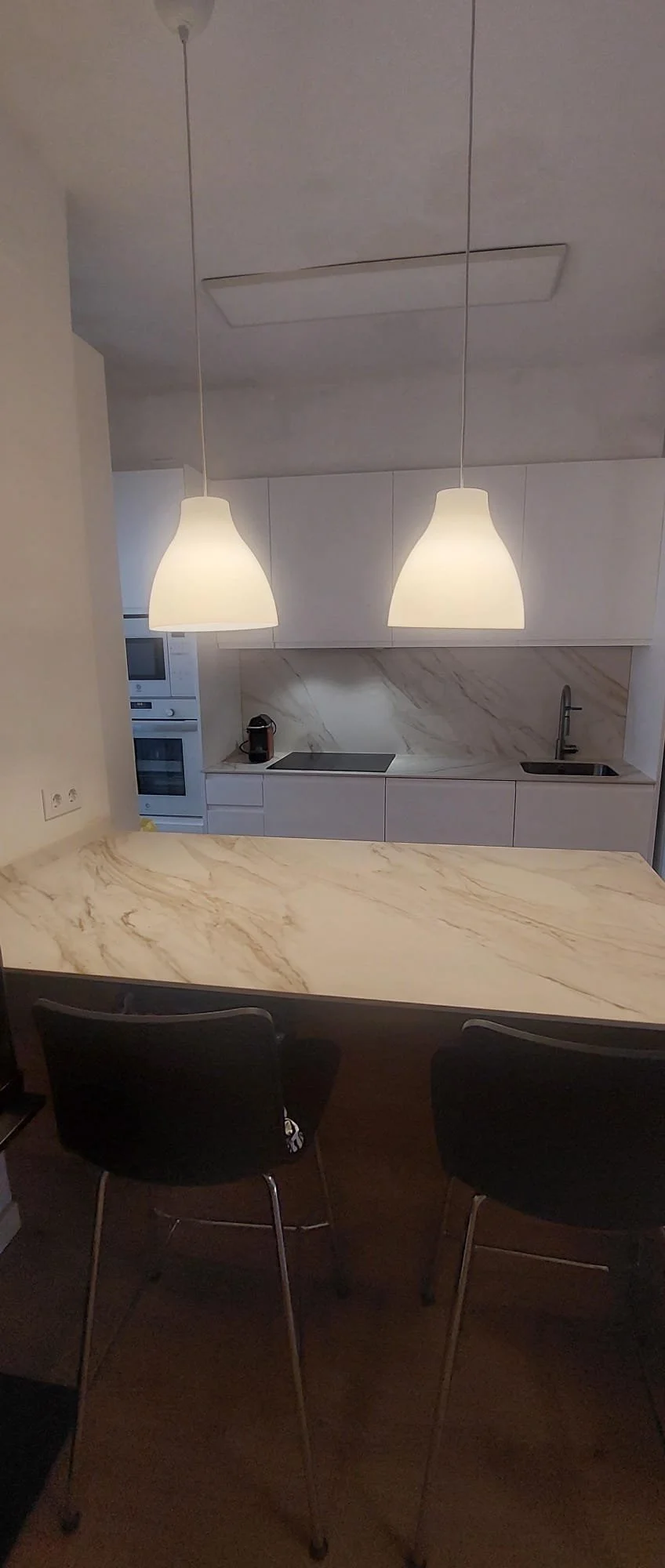 Cocina moderna con encimera de piedra beige, dos lámparas colgantes blancas, electrodomésticos integrados, fregadero y una cafetera encima de la encimera.