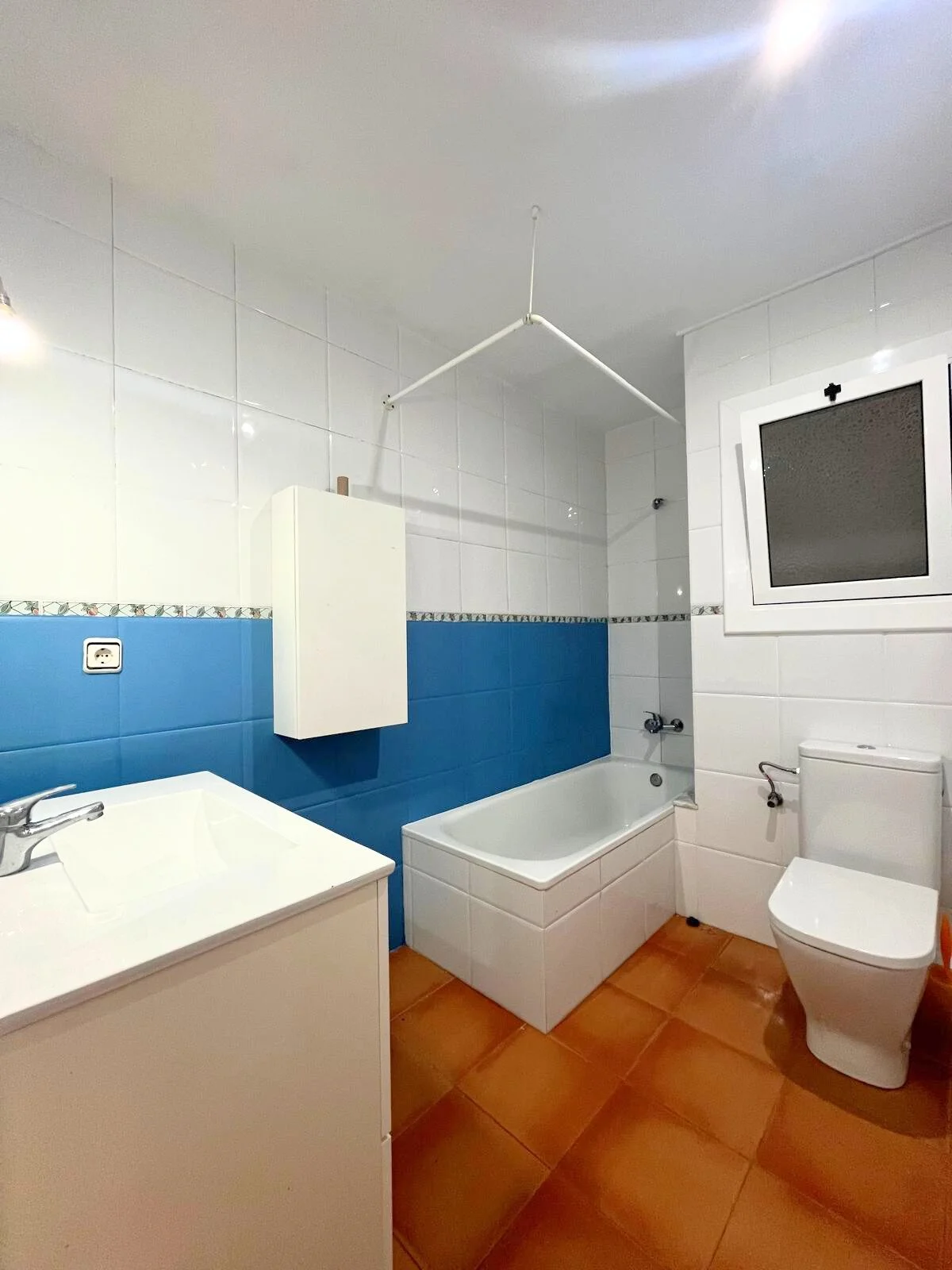 Baño con paredes blancas y azulejos azules en la mitad, bañera, inodoro y lavabo, ventana con marco blanco y suelo de baldosas marrones.
