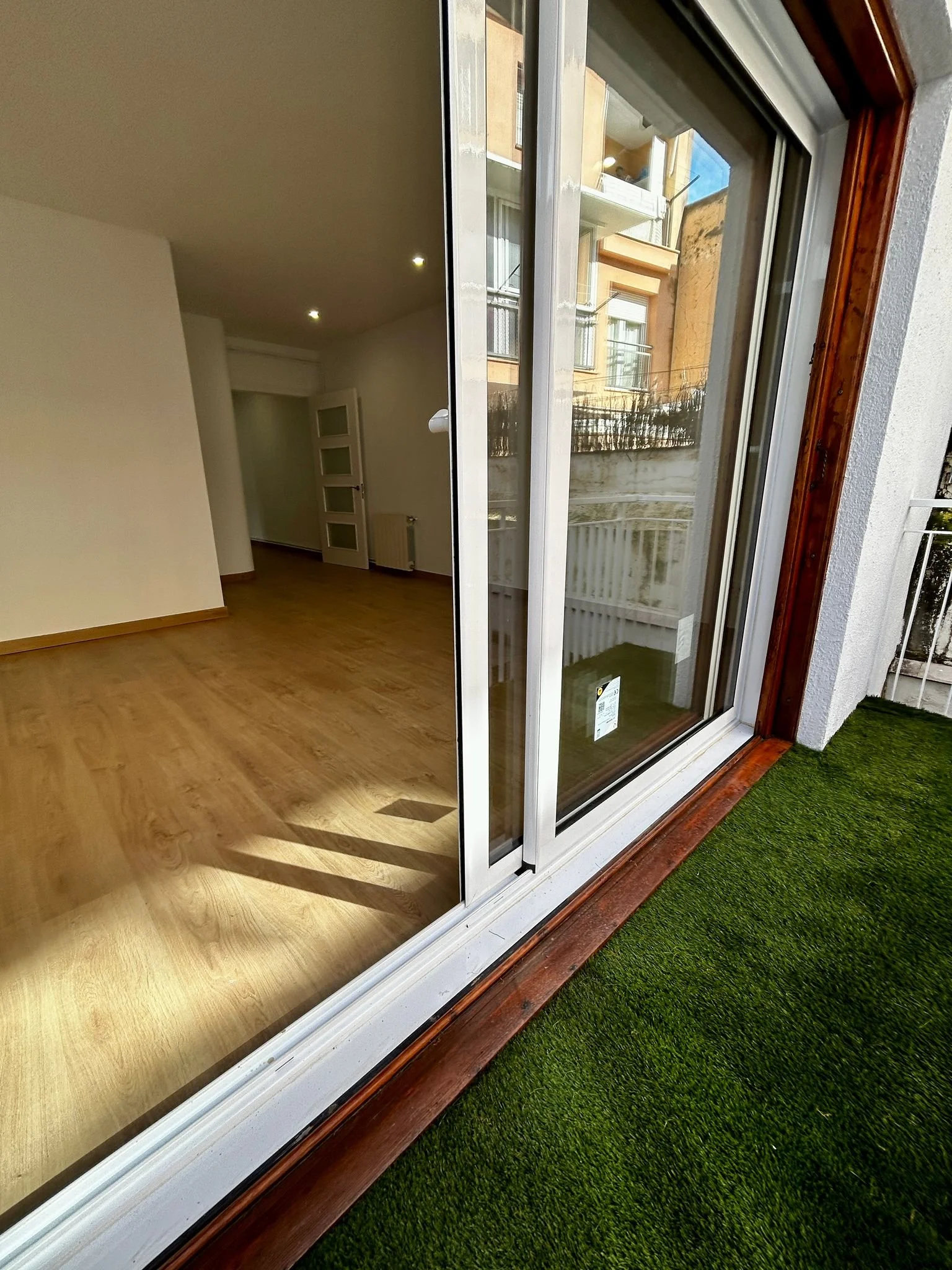 Vista desde un balcón con piso de césped artificial, puerta de vidrio con marco de madera que da a una sala vacía con pisos de madera y paredes blancas, y ventanas exteriores en edificios cercanos.