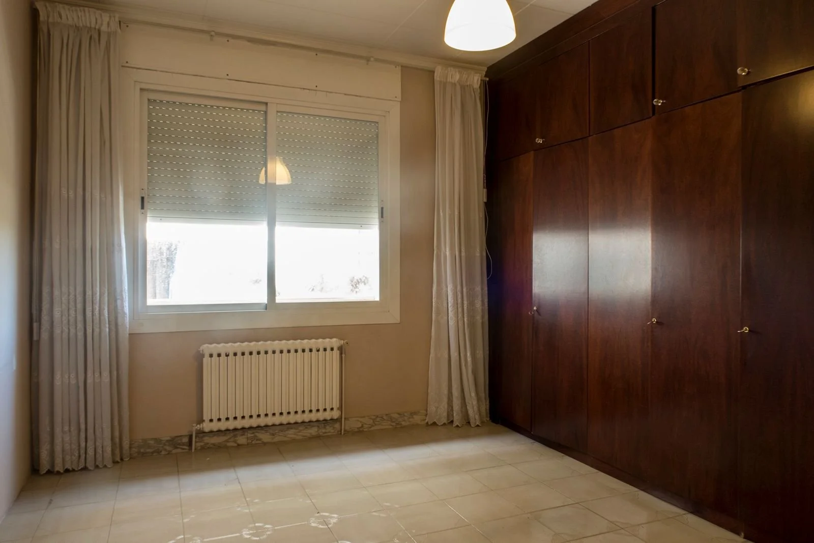 Habitación con ventana, cortinas beige, armario de madera oscura y una lámpara de techo.