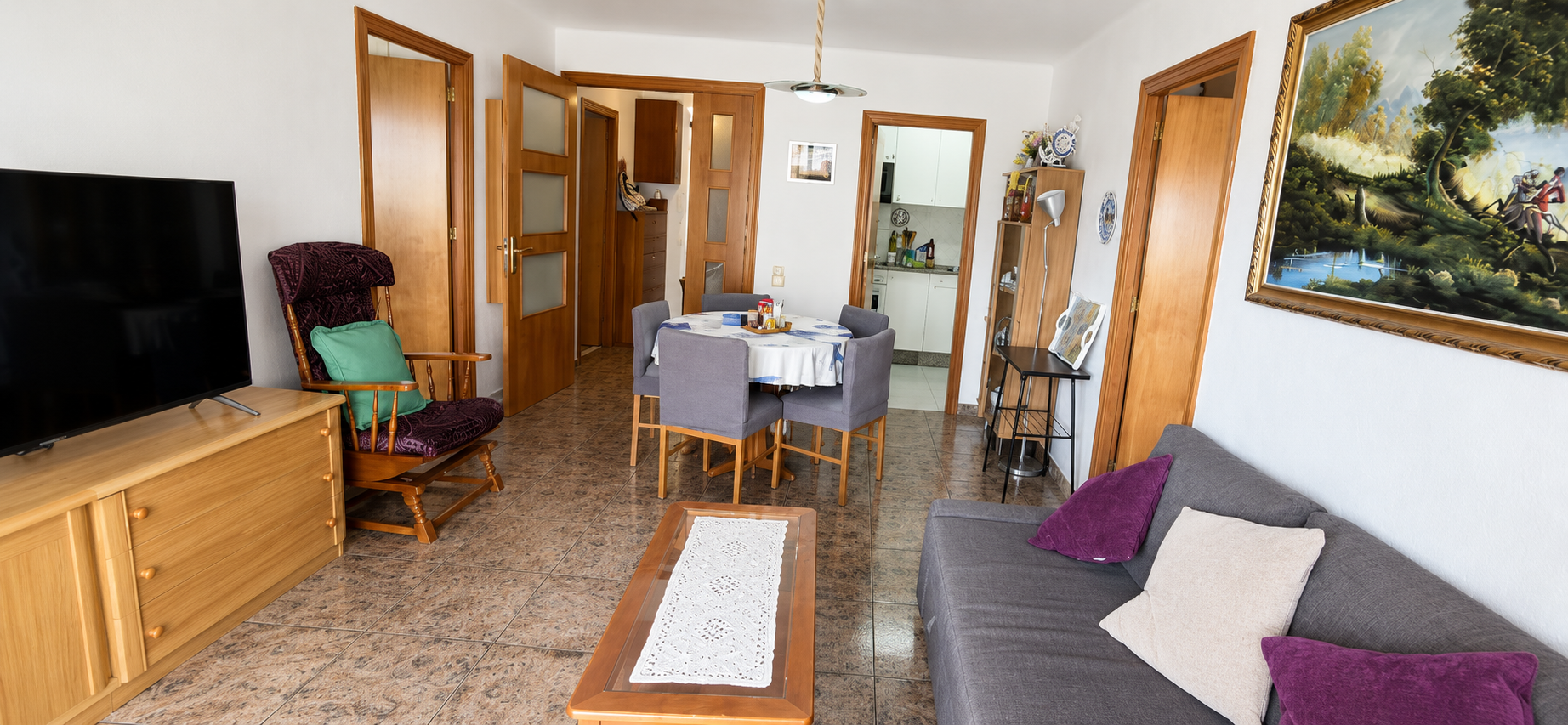 Sala de estar con sillón gris, sofá con cojines púrpuras y beige, mesa de madera, televisor, comedor con mesa y sillas, vista a la cocina, cuadros y decoración en paredes, madera en puertas y muebles, piso de baldosas.