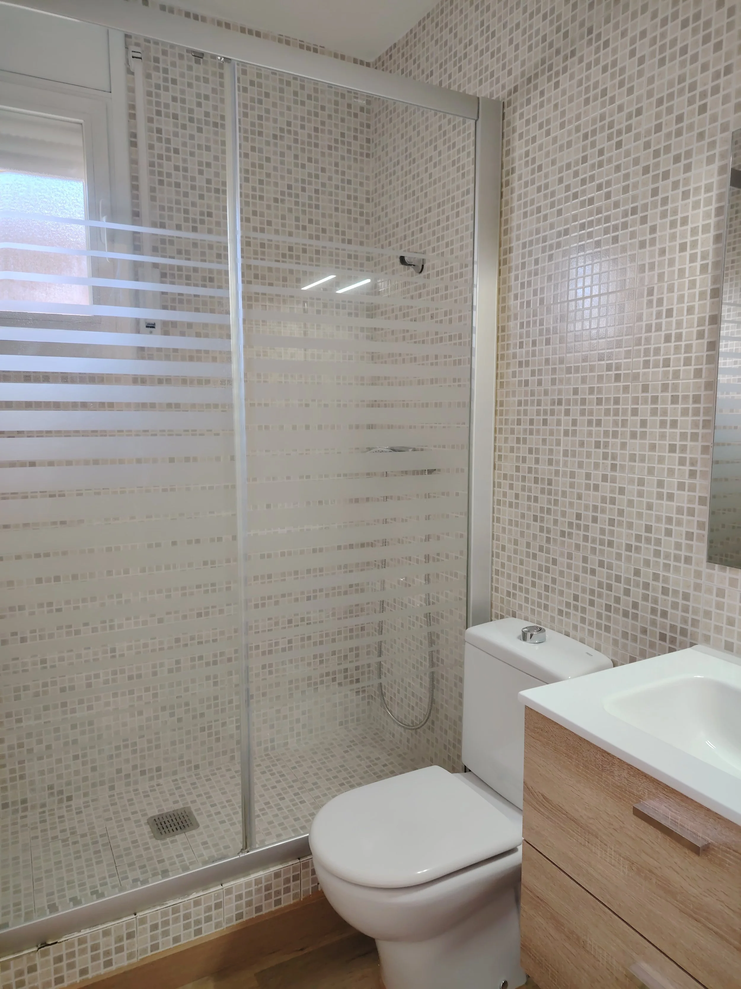 Baño con ducha con paredes de azulejos pequeños en tonos beige, inodoro y lavabo de madera.