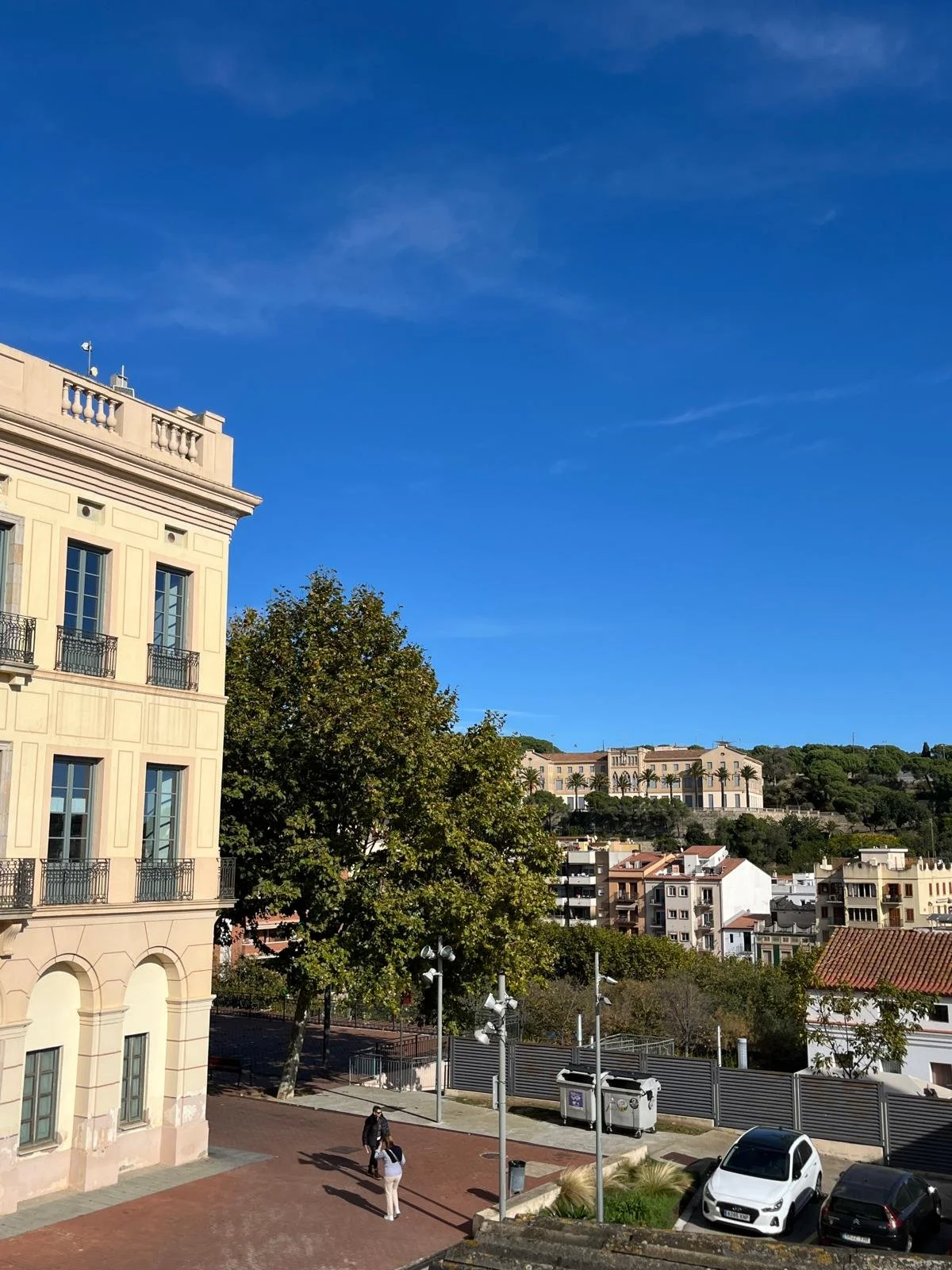 Escena urbana con edificio de estilo clásico, árboles y personas caminando en la calle, vehículos estacionados y un fondo de colinas con más edificios y vegetación, cielo despejado azul.