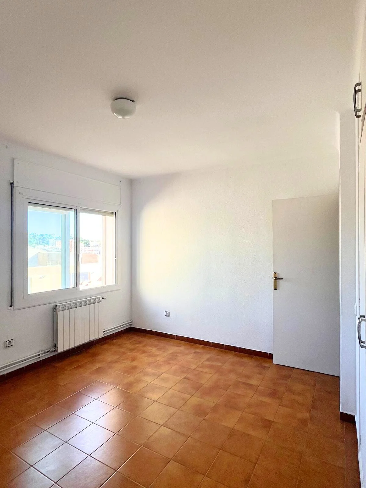 Habitación vacía con paredes blancas, piso de baldosas color terracota, ventana grande con vista a edificios, puerta blanca y radiador en la pared, luz natural en la habitación.