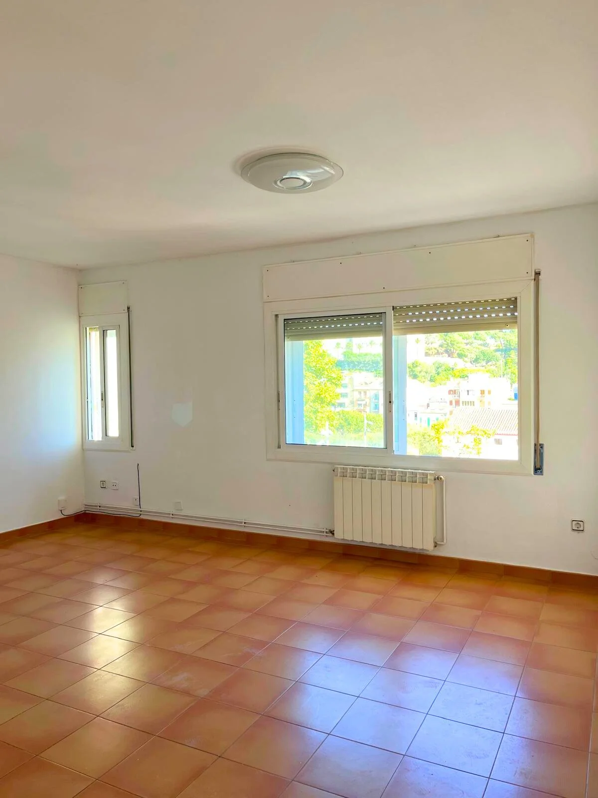 Sala vacía con paredes blancas, ventana grande con vista a edificios y árboles, piso de baldosas anaranjadas, radiador debajo de la ventana, y luminaria en el techo.