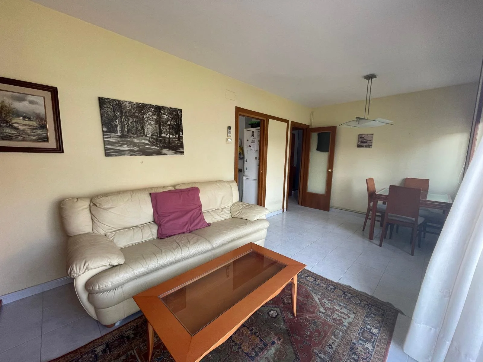 Sala de estar con sofá beige, mesa de madera con cristal, cuadros en la pared y comedor de madera con cuatro sillas. La habitación tiene puertas de madera y cortinas blancas.
