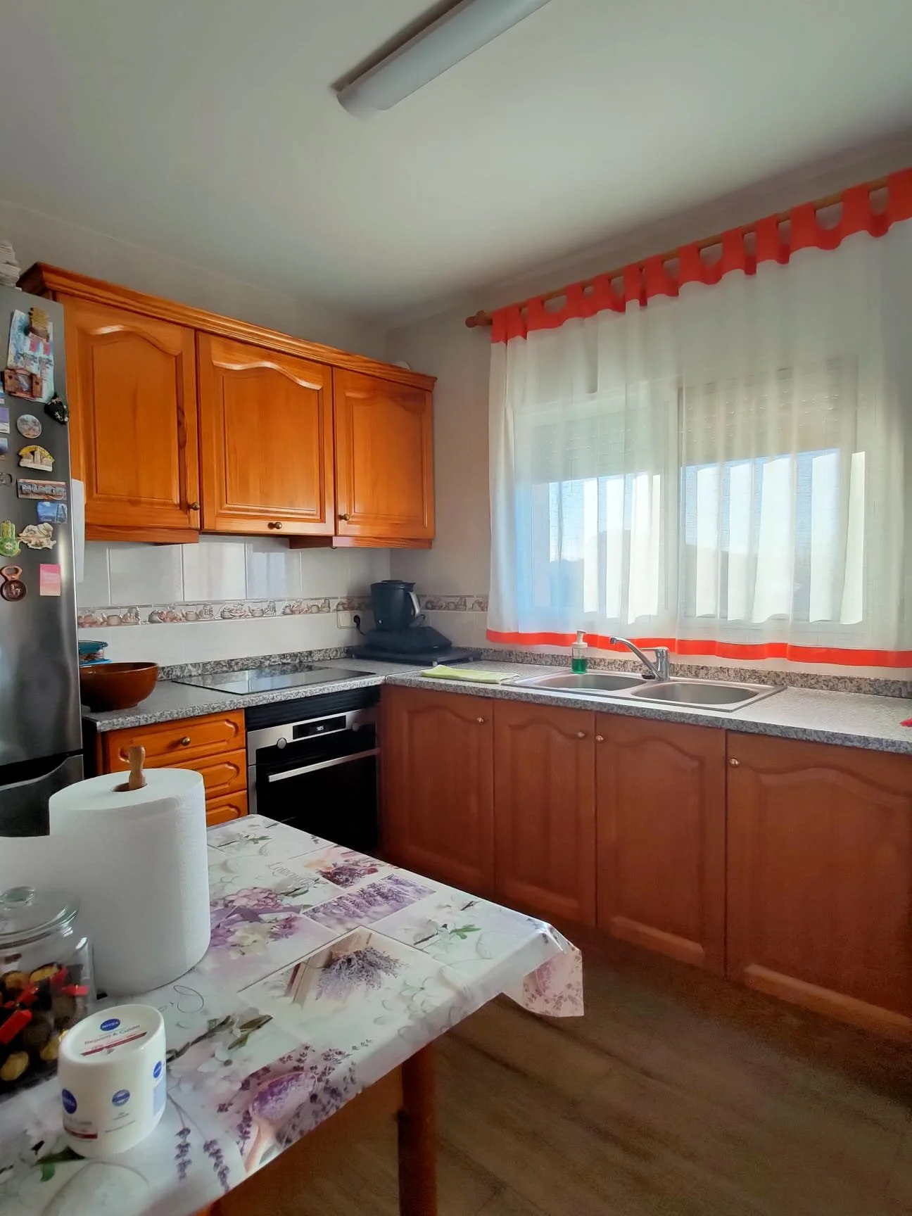 Cocina con armarios de madera, encimera de granito, ventana con cortinas blancas con borde naranja, mesa con mantel de flores y papel toalla, electrodomésticos y utensilios en la encimera.