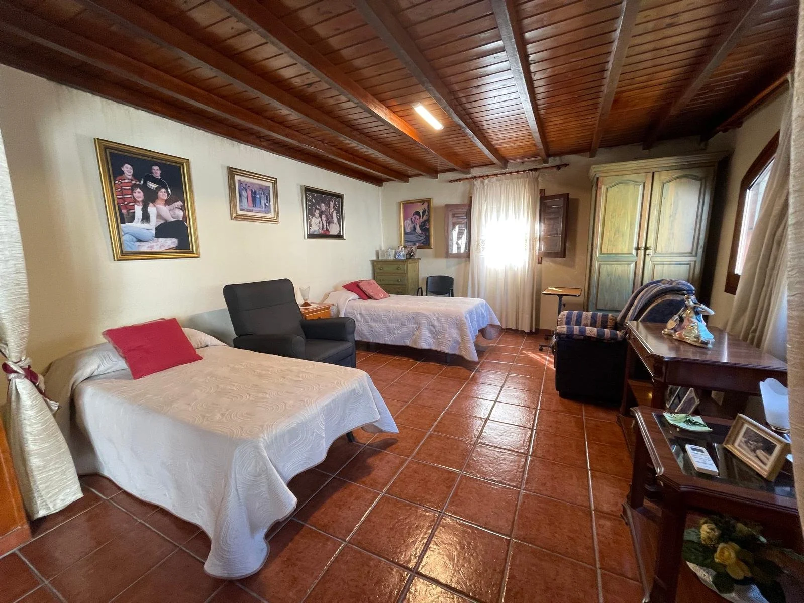 Una habitación con dos camas, una con cobertor blanco y otra con cobertor beige, varias sillitas, un armario grande, cuadros en la pared, y ventanas con cortinas blancas, en un entorno de madera y cerámica. Casa en Malgrat de mar.