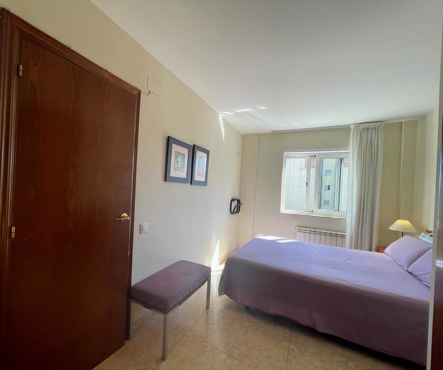 Habitación con cama, ventana, cortinas blancas, armario grande de madera y un banco con tapete