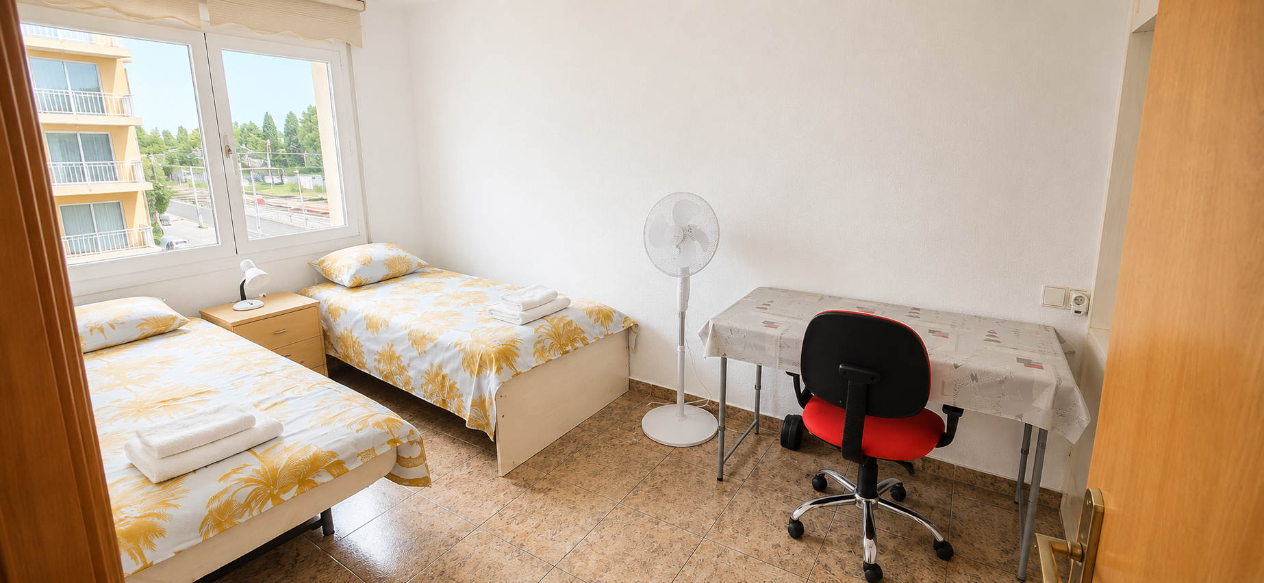 Habitación con dos camas, vista a un edificio de apartamentos desde la ventana, escritorio con silla, ventilador y lámpara de mesa.