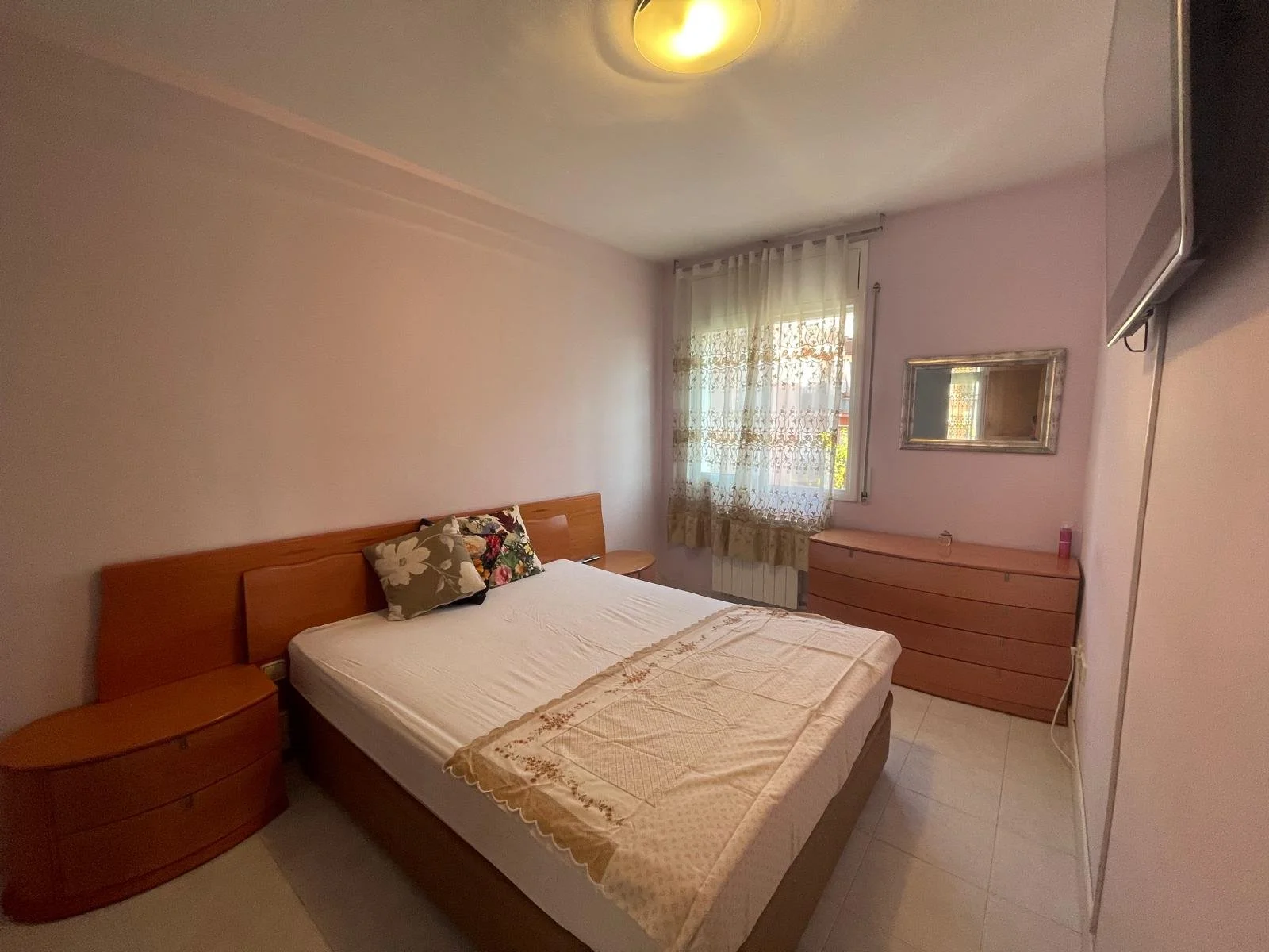 Habitación con cama doble, una almohada con funda floral, cortinas translúcidas en la ventana y muebles de madera clara, incluyendo una consola y mesitas de noche.