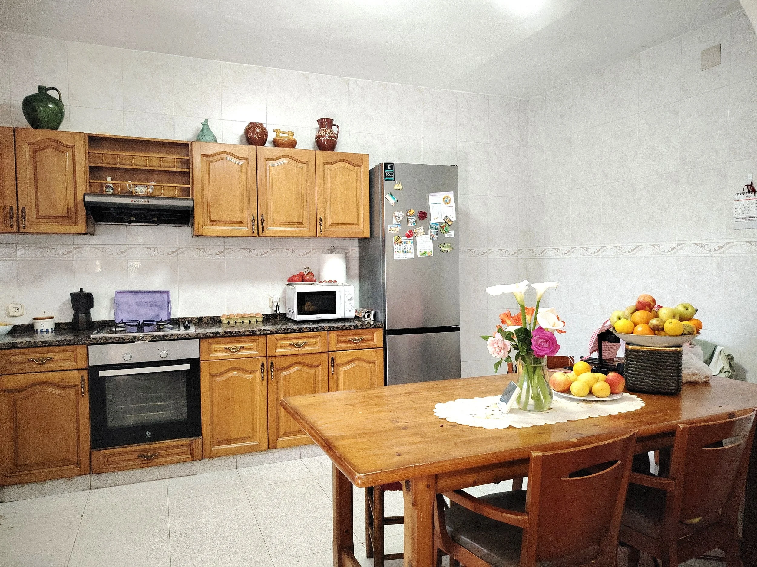 Cocina con gabinetes de madera, una estufa, microondas y nevera, mesa de madera con flores y frutas, y paredes de azulejo claro.