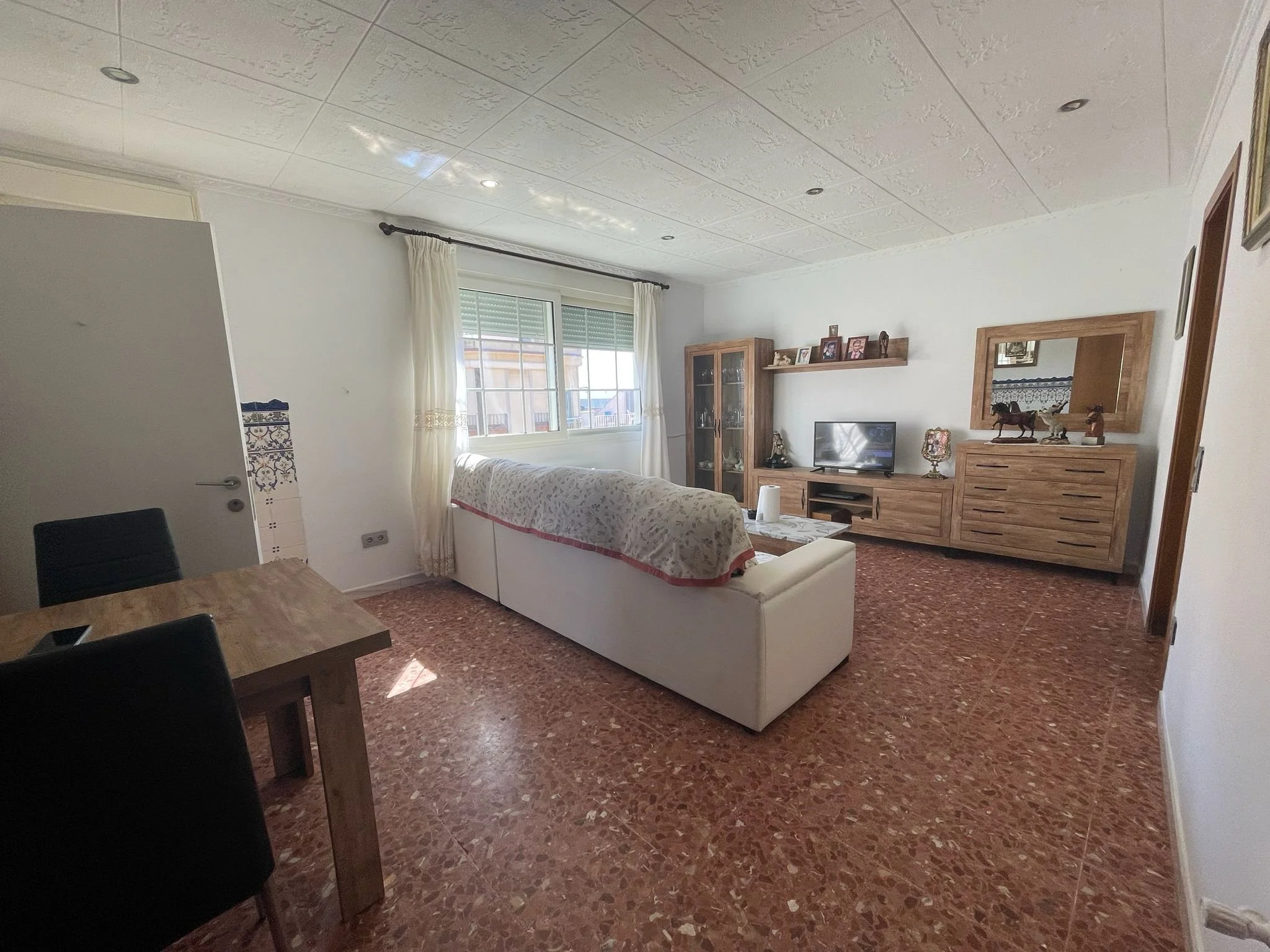 Sala de estar con sillones, un mueble de madera con cuadros y decoraciones, y una ventana con cortinas claras.