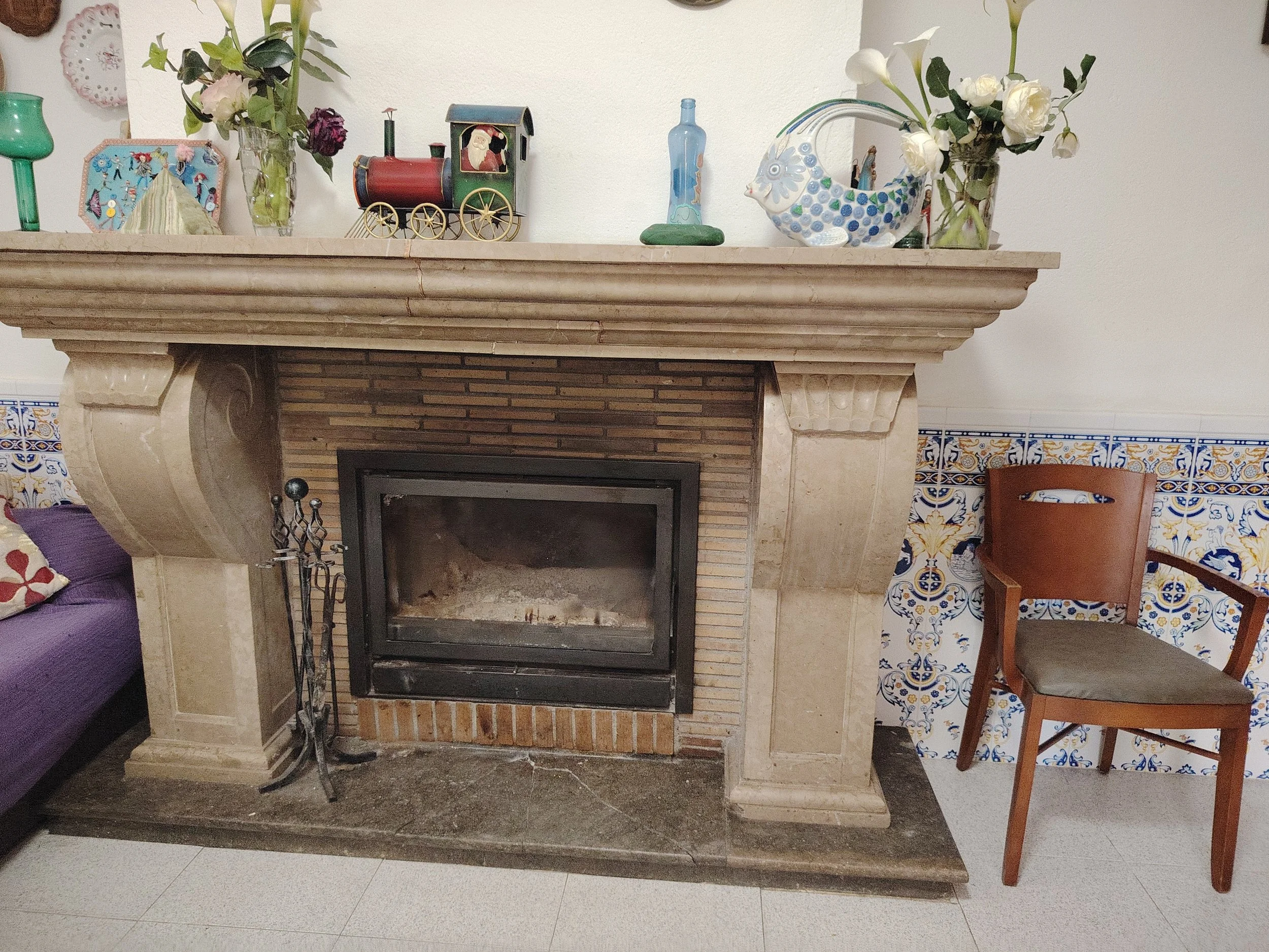 Una repisa de madera con decoraciones variadas, incluyendo un reloj, un vaso con flores, una figura de tren con Santa Claus, un pájaro decorativo de cerámica, y más plantas y objetos decorativos en el fondo. La repisa está sobre una chimenea de piedra con mosaicos azules y dorados.