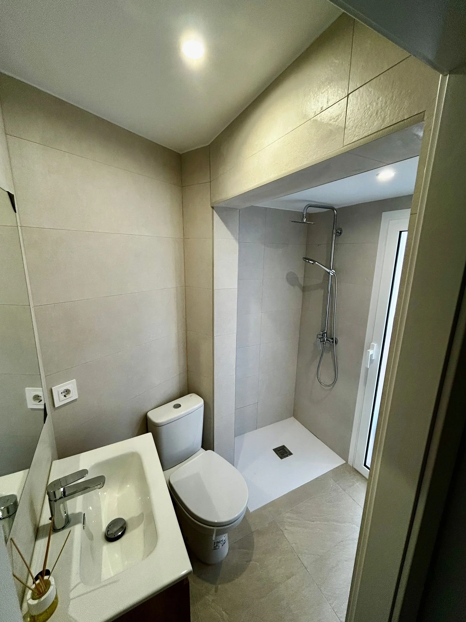 Baño moderno con lavabo, inodoro y ducha, paredes y suelo en tonos beige, ventana y luces empotradas en el techo.