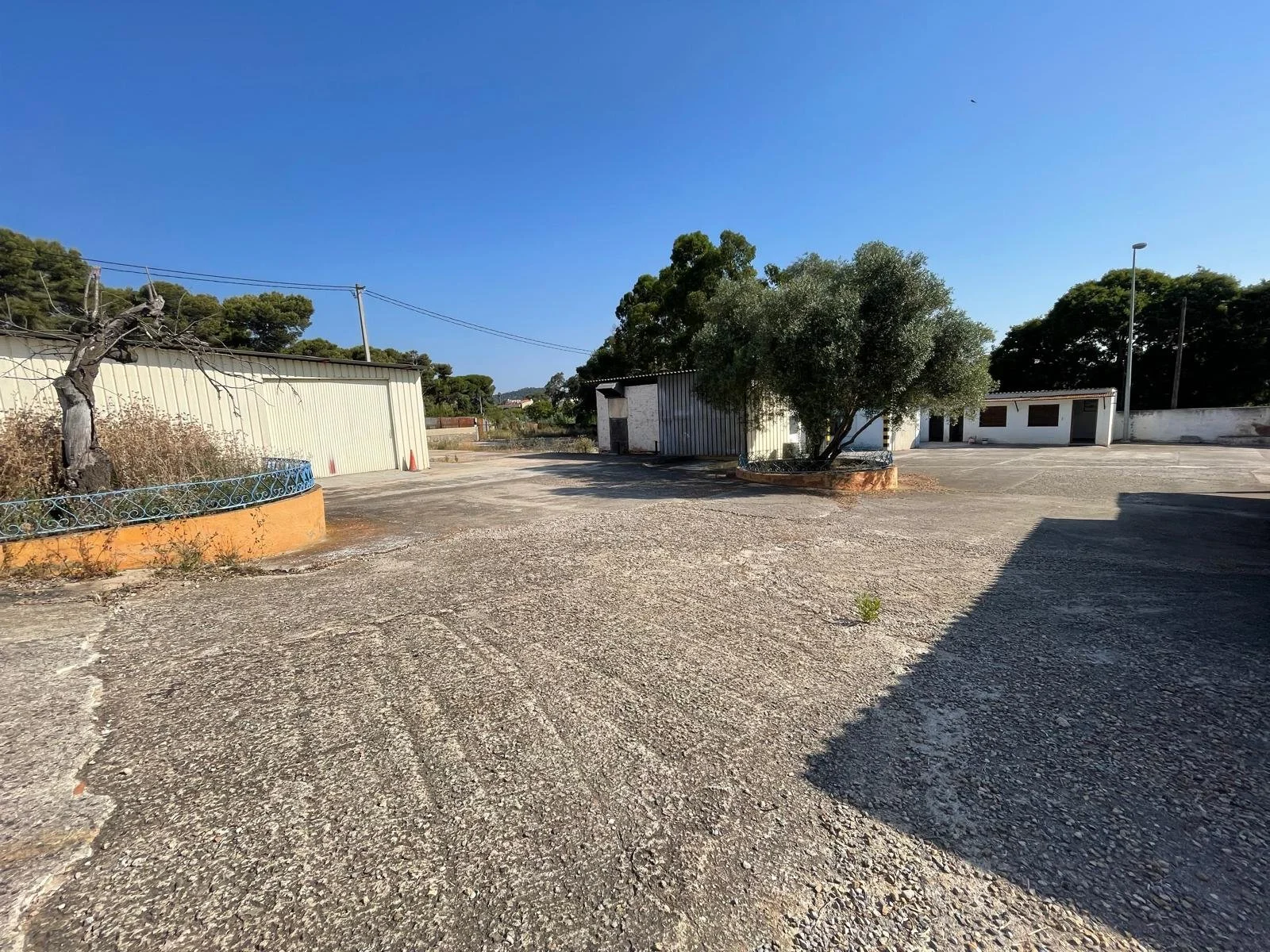 Un espacio abierto con suelo de grava, árboles y estructuras de color blanco y gris bajo un cielo despejado y azul. Casa en Malgrat de mar.