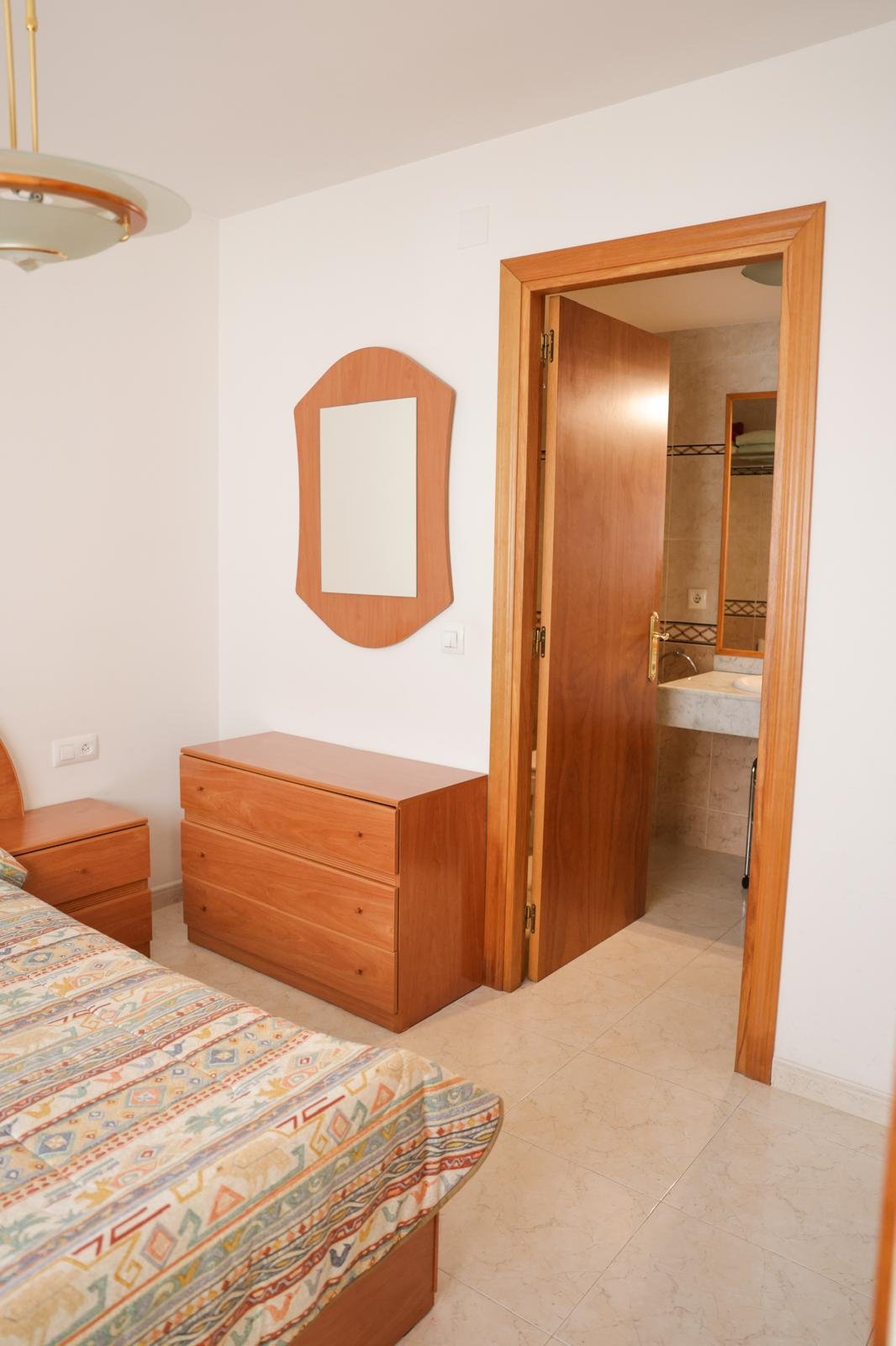 Imagen de una habitación con un espejo de madera, una cómoda también de madera, parcialmente visible una cama con colcha colorida y un baño con lavabo visible desde la puerta entreabierta.