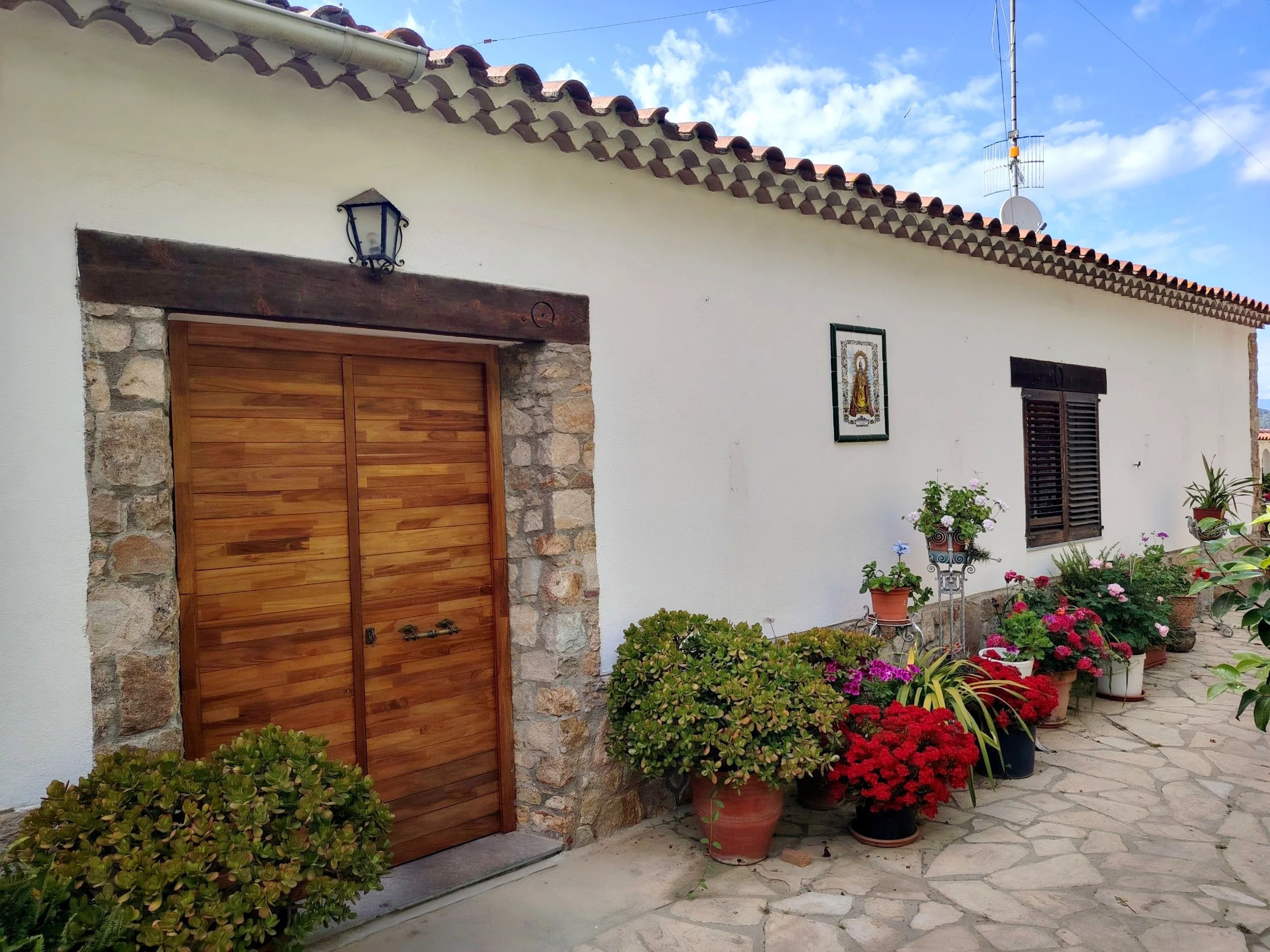 Casa con paredes blancas, tejado de tejas rojas, puertas de madera, paredes de piedra, jardín con muchas plantas y flores en macetas, dibujo religioso en la pared, cielo con nubes.